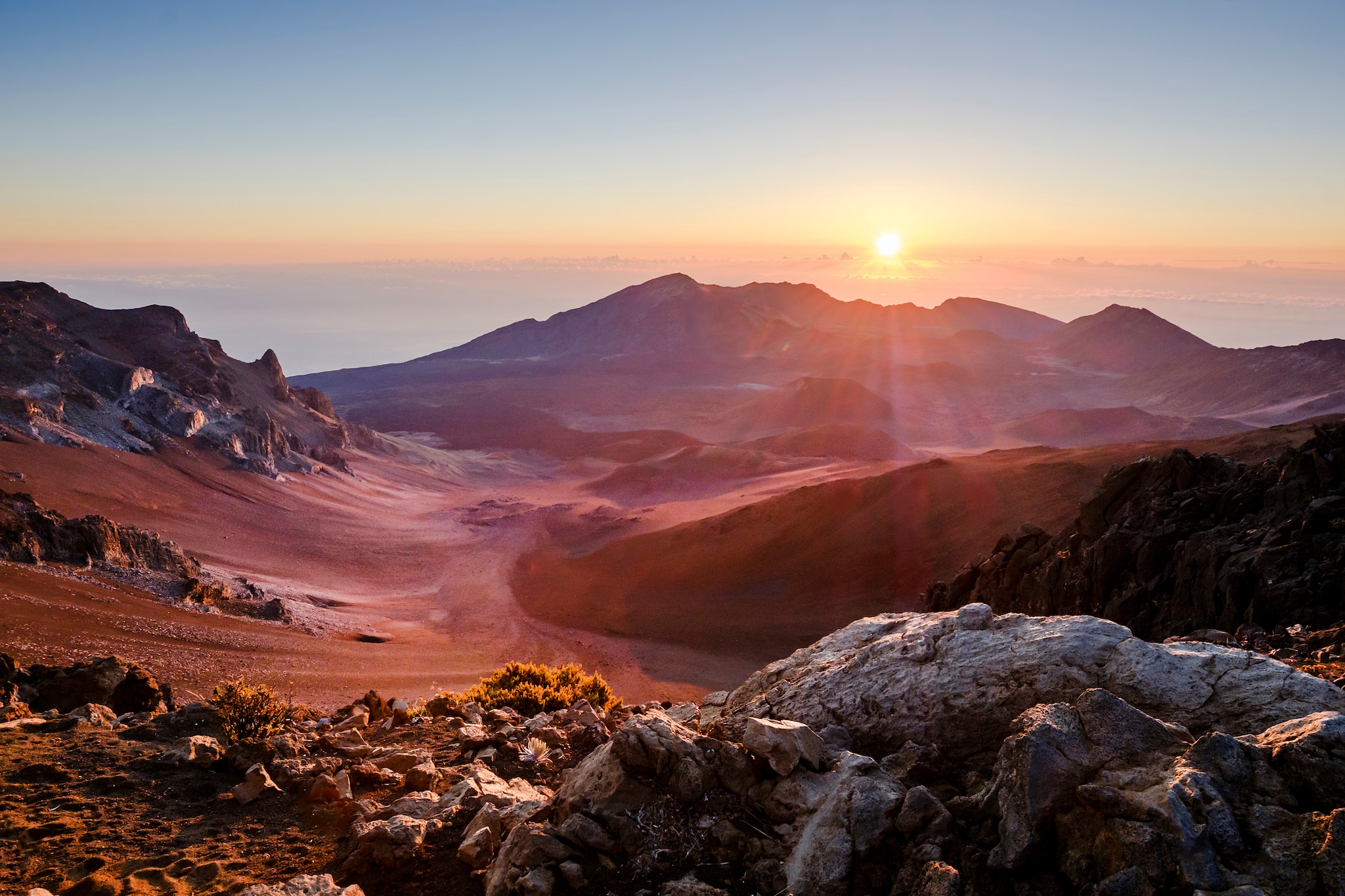 At sunrise, the Haleakala Crater is unearthly, with red sand and low-growing shrubs scattered across rugged volcanic rock.
