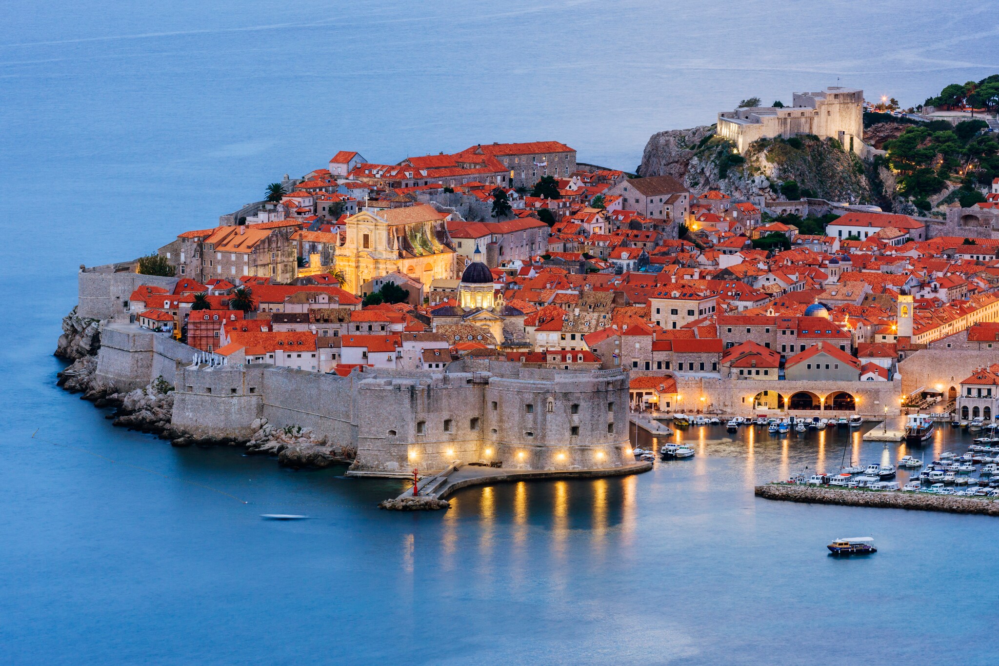 Aerial view of Dubrovnik’s Old Town, tightly clustered buildings with tan stone and red roofs separated from the Adriatic Sea by tall walls.