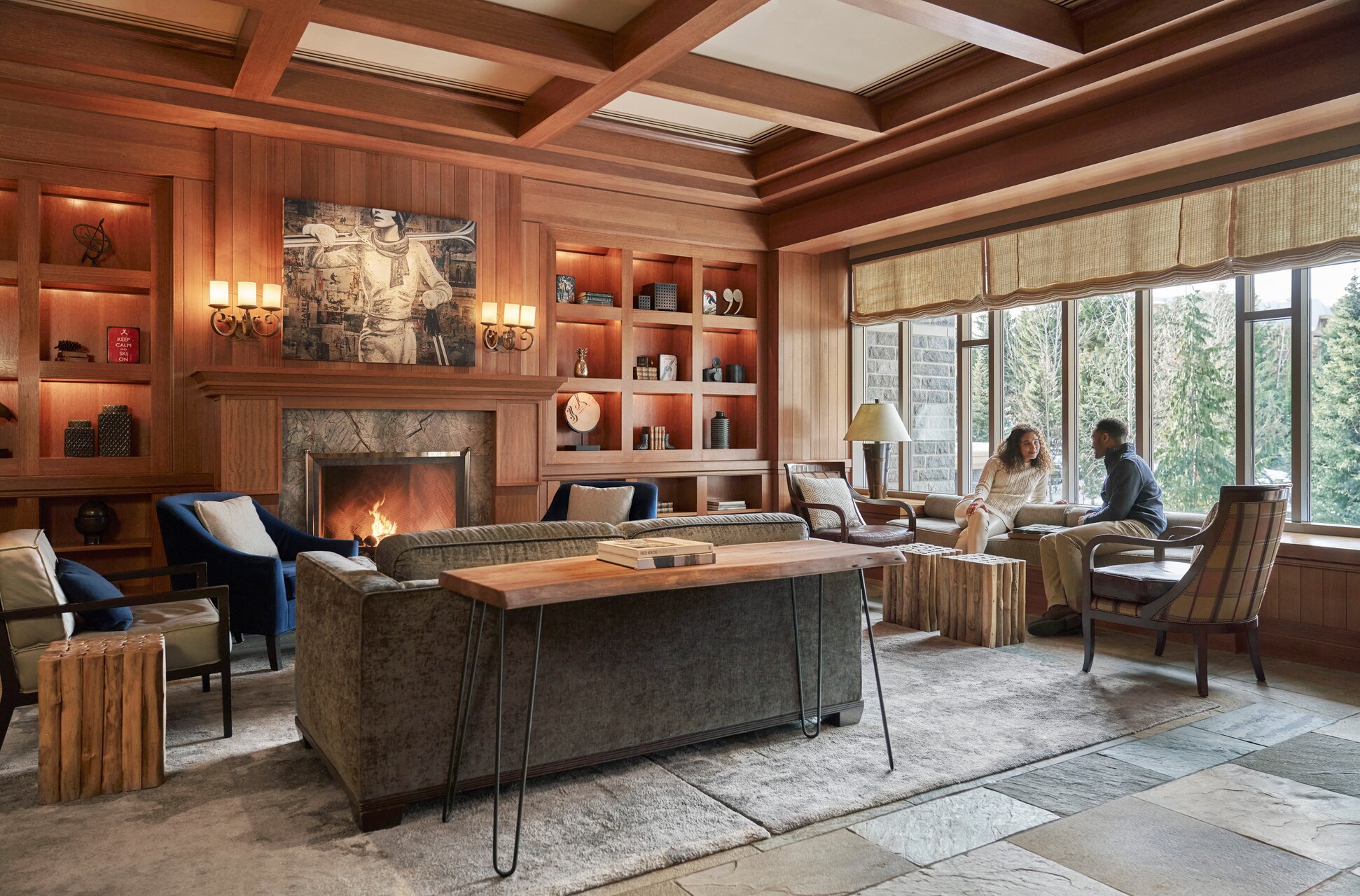 In a lodge-like seating area, a Black couple lounges on window seats while a fire roars under a painting of a woman in ski gear.