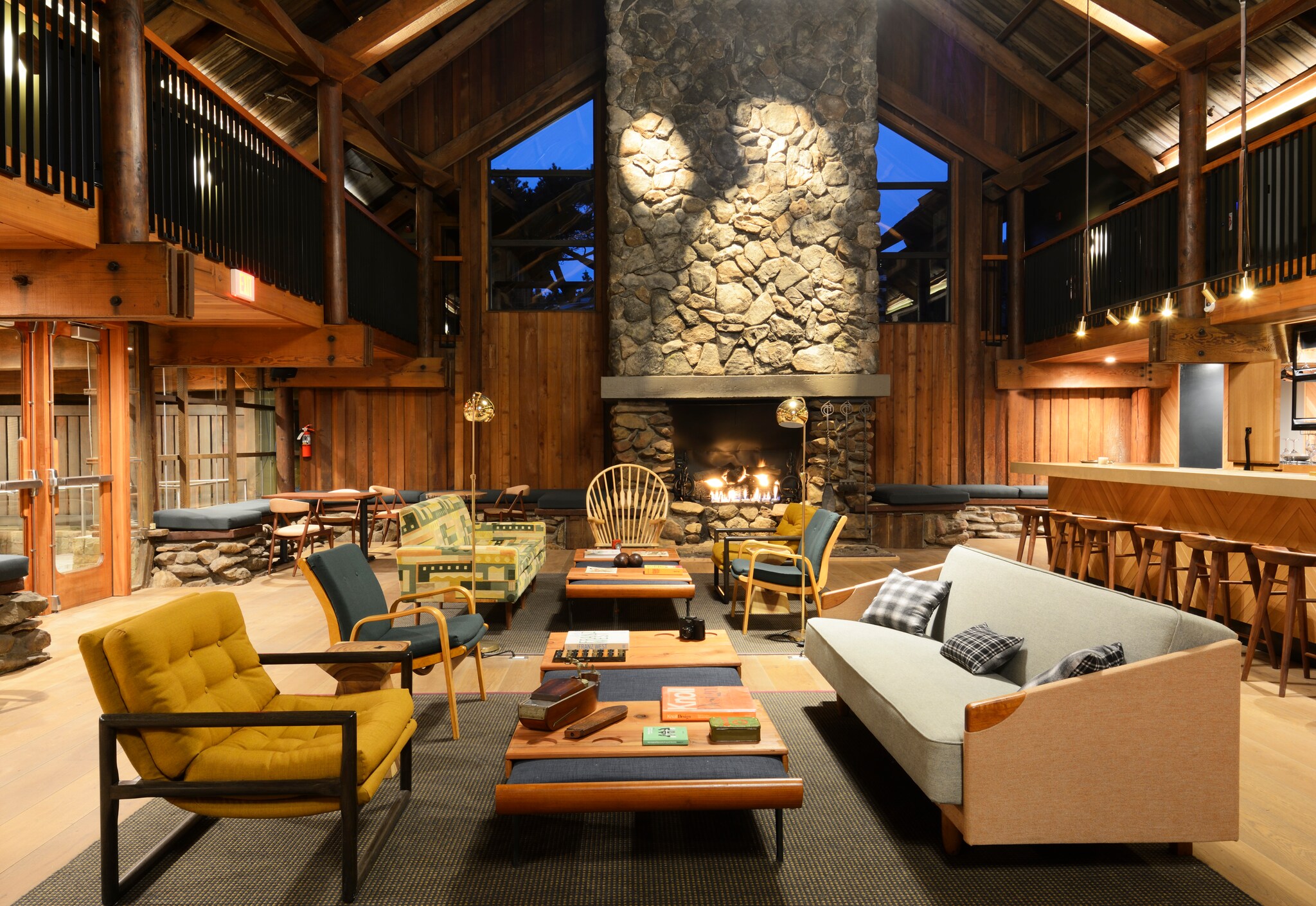 The hotel lobby features simple furniture in browns and yellows under a high, gabled ceiling. A stone fireplace dominates the far wall.
