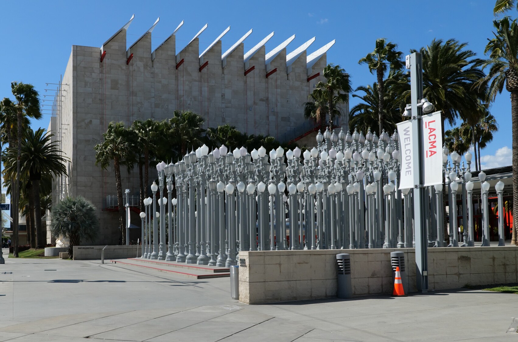 An installation of rows and rows of silver street lamps fills a courtyard in front of the museum, a tall gray building with a serrated roof.