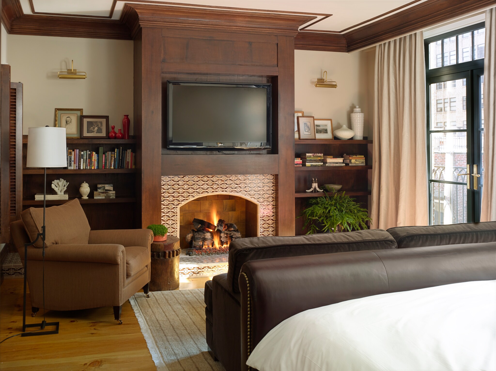The Master Bedroom of the North Moore Penthouse Suite has a large bed, a roaring fire under a mounted TV, and a plush armchair.
