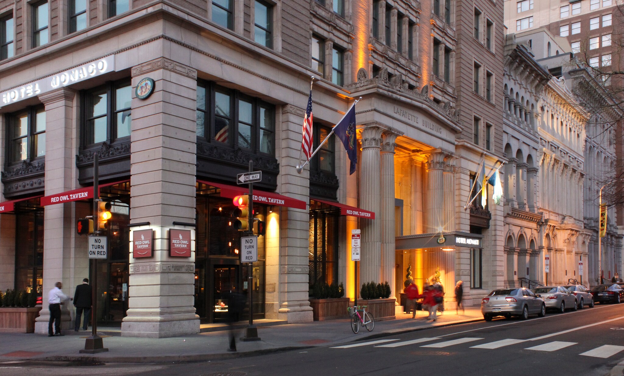 Exterior of Kimpton Hotel Monaco Philadelphia, a neoclassical building on the corner of a block, with red signs advertising Red Owl Tavern.