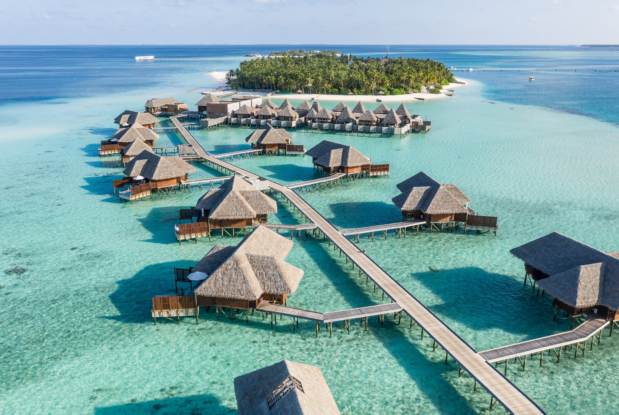 An aerial view of the Conrad Maldives Rangali Island shows multiple standalone overwater bungalows atop a calm blue ocean with a small and lush island in the background.