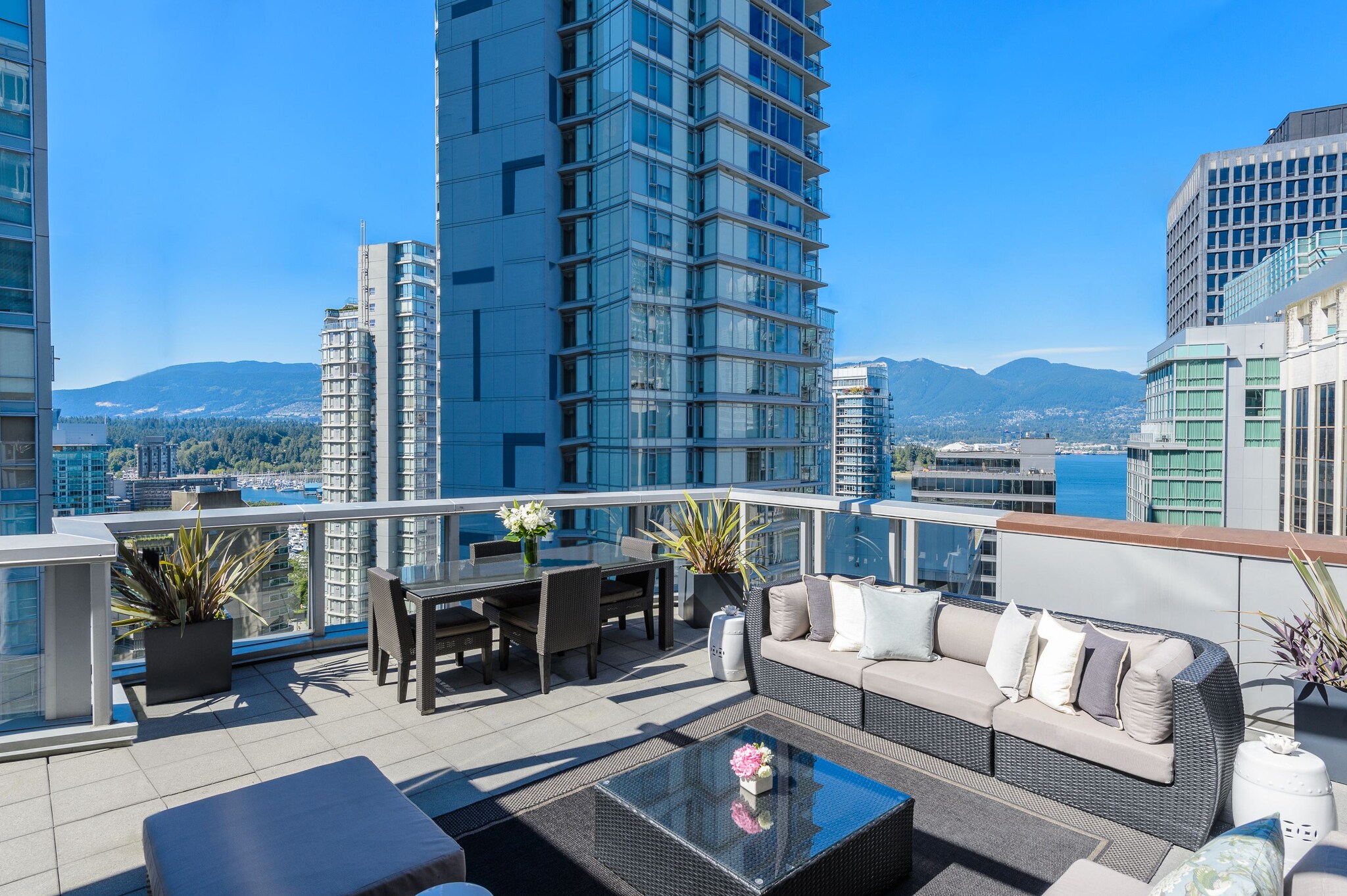 An elevated outdoor patio is furnished with a dining table and gray wicker couch. Over the railing, skyscrapers give way to water and mountains.