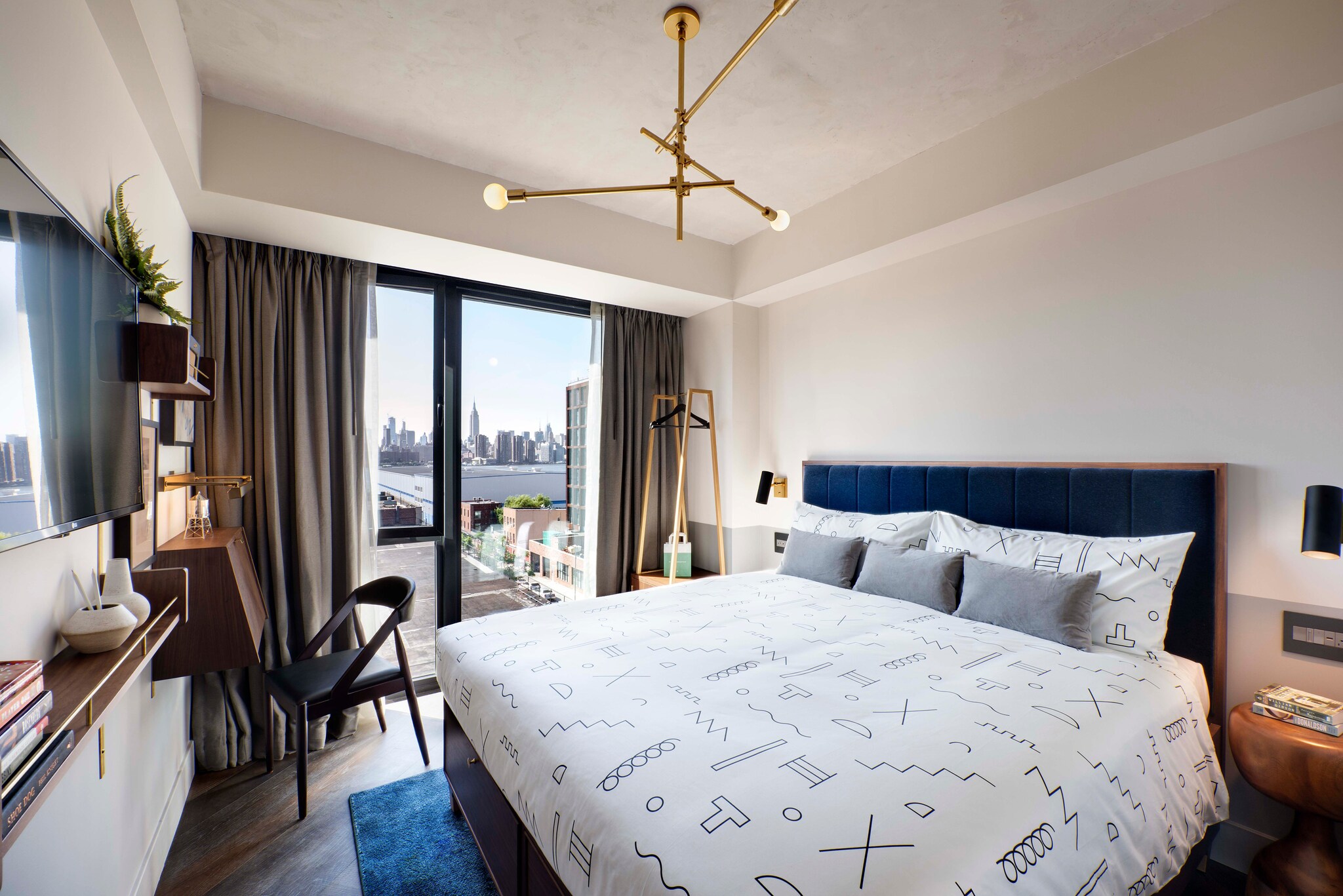 A guestroom has a large bed with a blue headboard and white bedding covered in neat scribbles. Floor-to-ceiling windows overlook Manhattan.