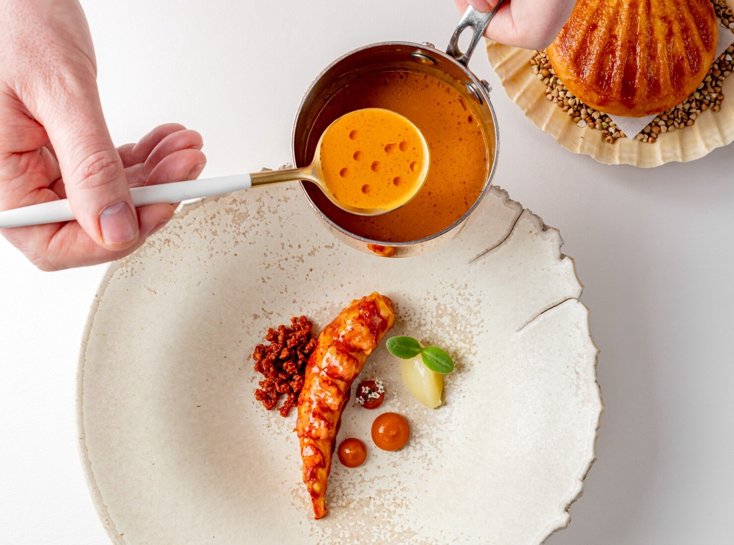 A white chef’s hand spoons orange sauce over a lobster tail garnished with a small sprout, a red crumble, and dollops of red sauce.