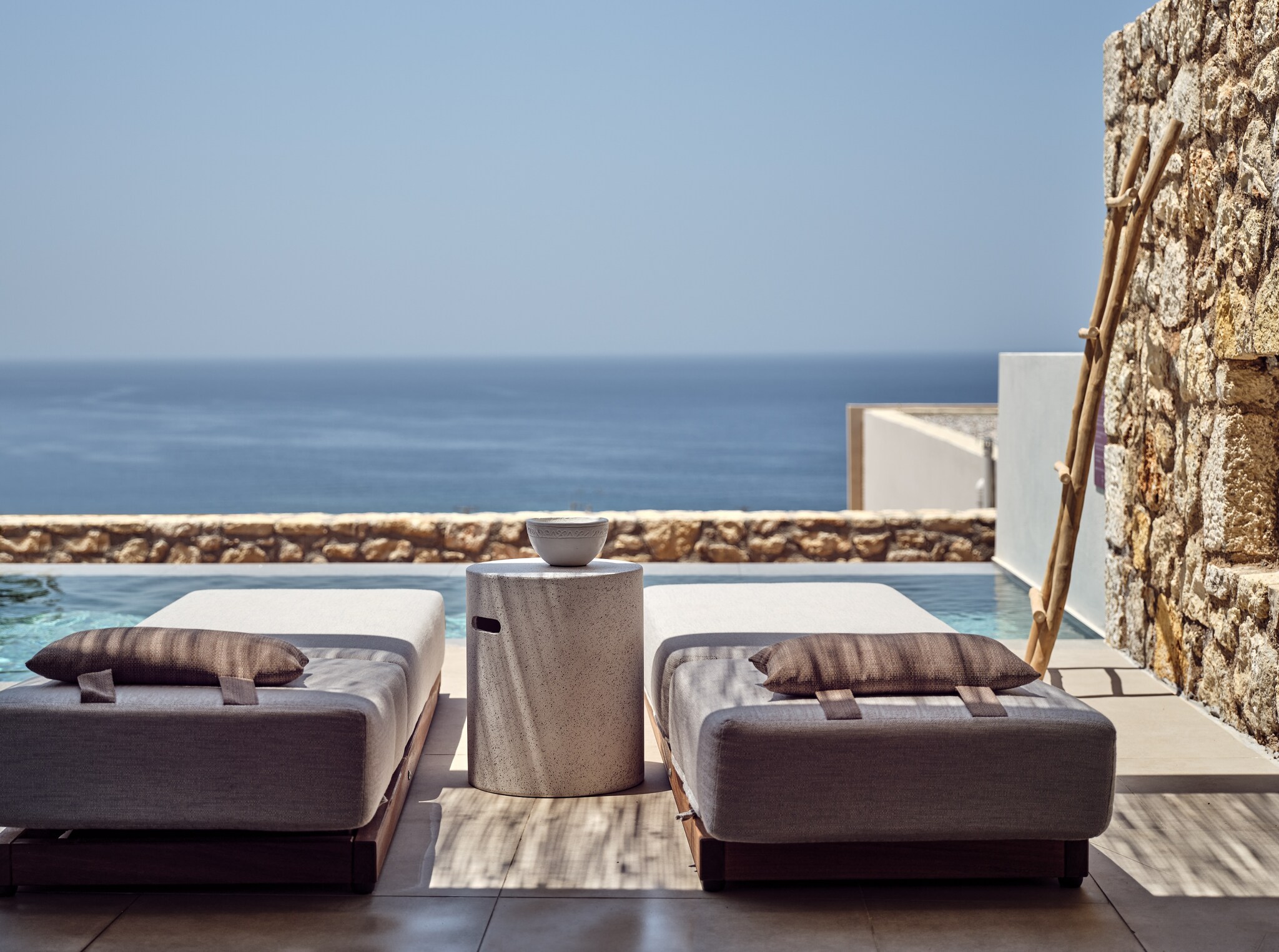 Two flat daybeds are arranged in front of a small rectangular pool, which is separated from the sea by a low rock wall.