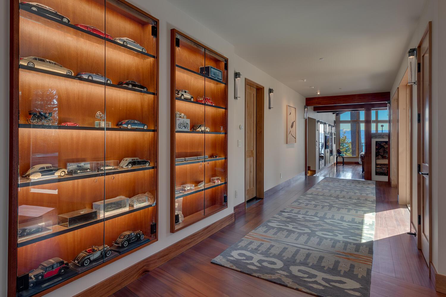 A long, wide hallway is set with a gray patterned rug, and vintage model cars line backlit shelves covered by glass doors.