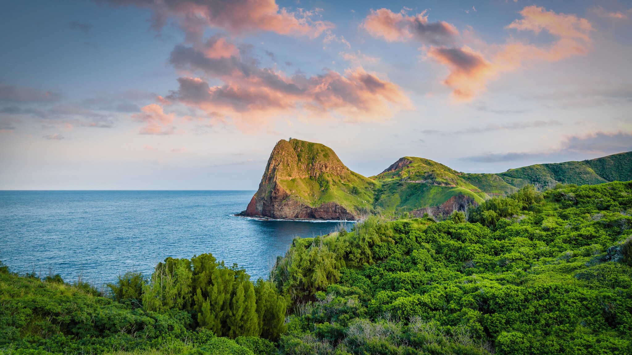 View from afar of Kahakuloa Head at sunset. On the promontory, lush greenery gives way to brown oceanside cliffs.