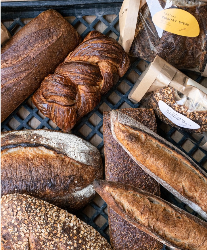 Overhead view of bread on a blue cooling grid, baguettes, braided bread, and seedy bread ready for sale.
