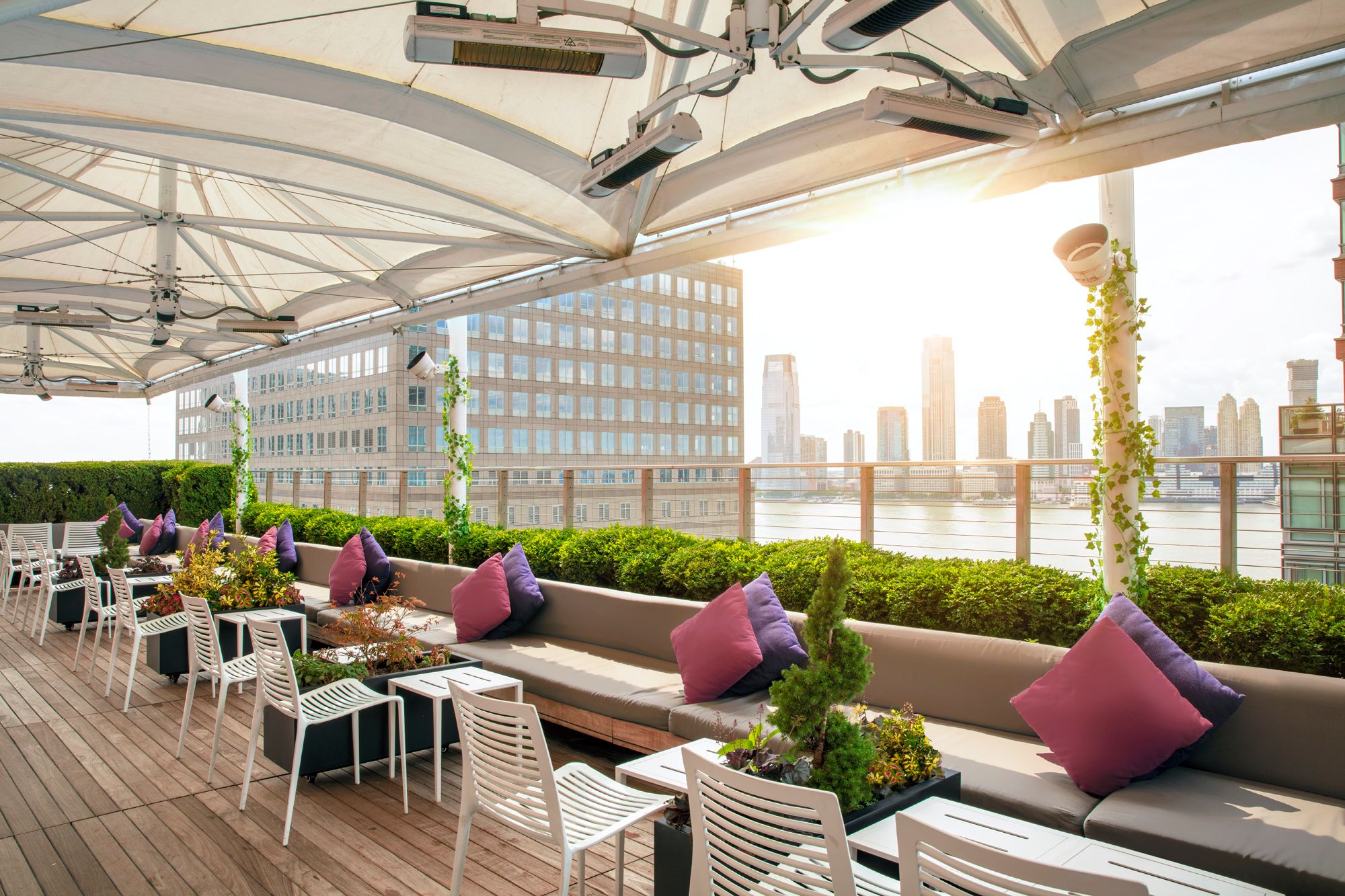 In the daytime, the Loopy Doopy Rooftop Bar looks across the bright water beyond. Lush bushes grow up behind dining couches.