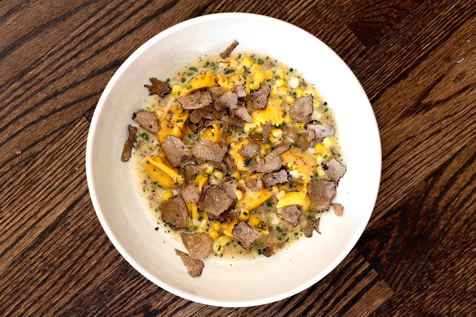 Overhead view of a large white dish of agnolotti in a sauce filled with corn and herbs, topped with fine shavings of black truffle.