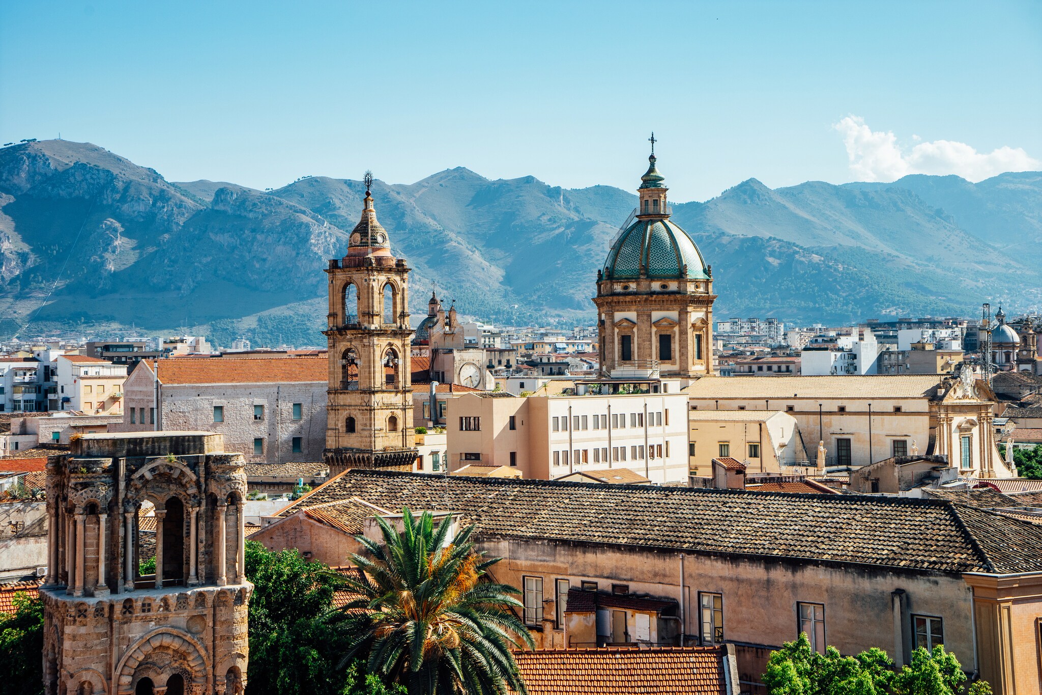 View over the terracotta roofs and cathedral domes of Palermo, with craggy hills beyond.