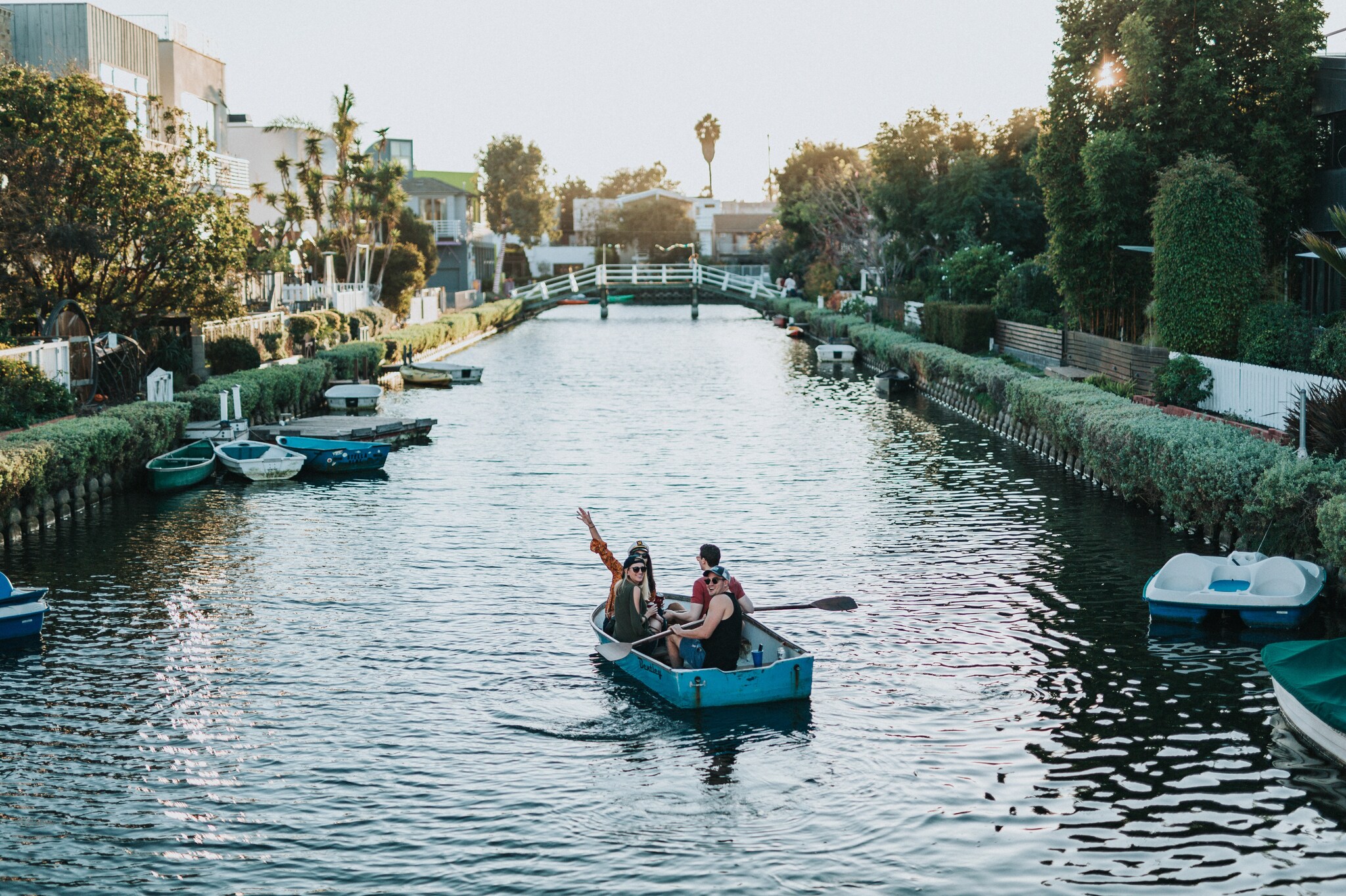 Four adults are crowded into a blue rowboat, traveling down a canal lined by low hedges and small trees.