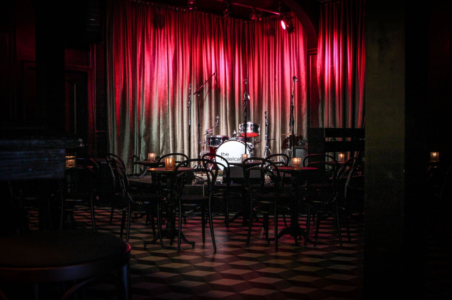 Tables and chairs are silhouetted in a dark room against a stage with a drum set, microphones, and red spotlights on a silver curtain.