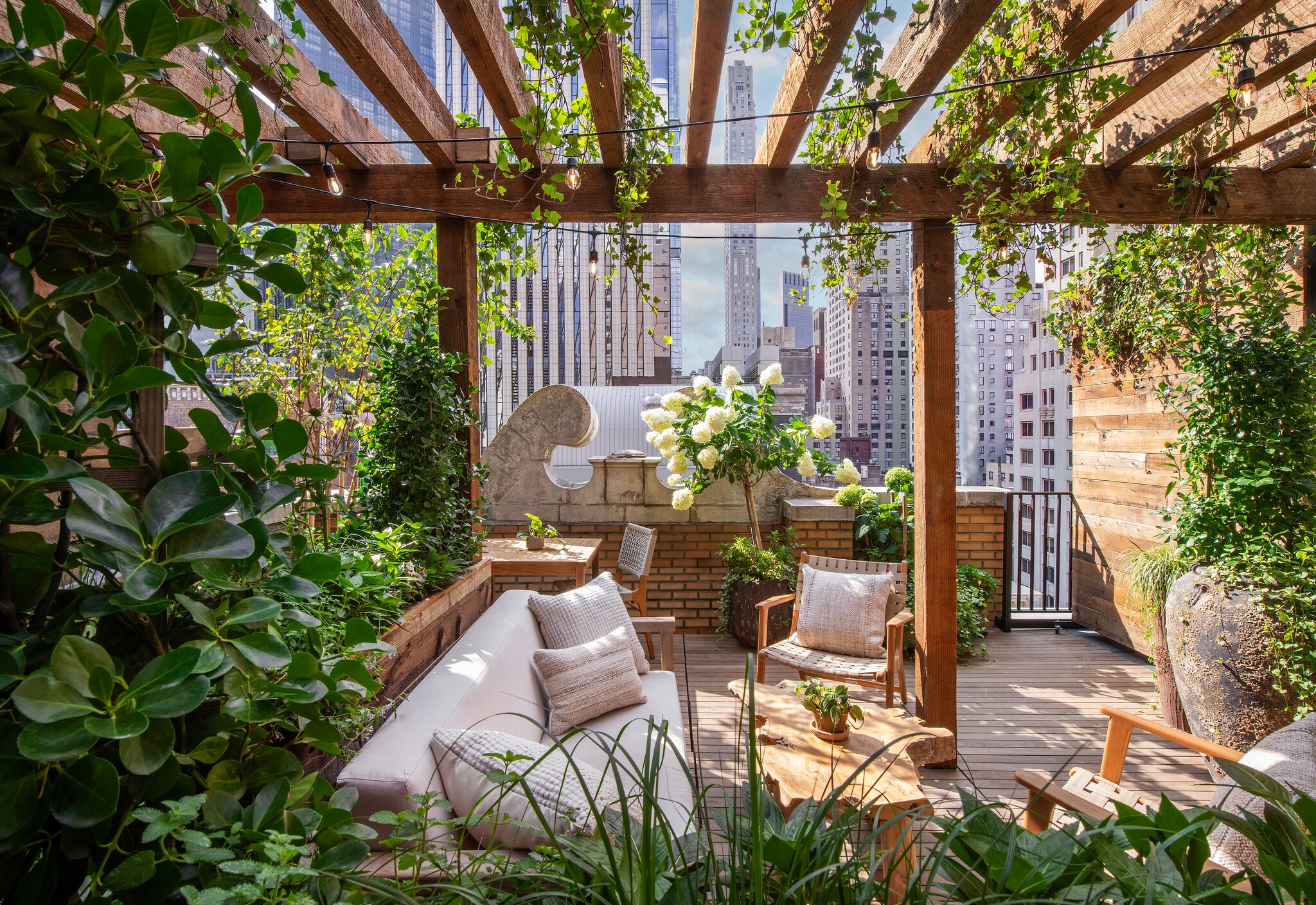 The space outside the Pergola Terrace King is overgrown with vines and lush potted plants. A couch and chairs surround a coffee table.