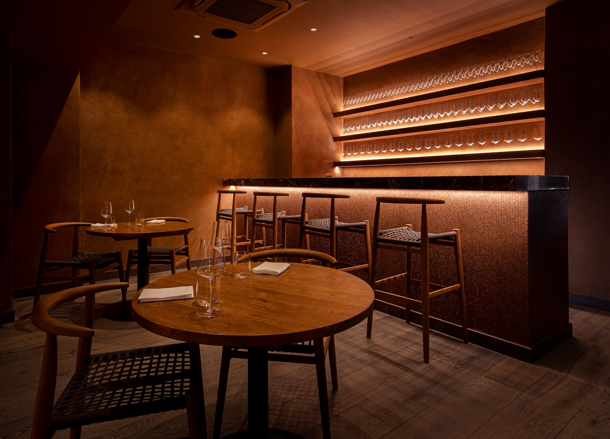 The interior of Akoko near the bar is dim, with red-brown walls, wooden tables and chairs, and slim, modern furniture profiles.