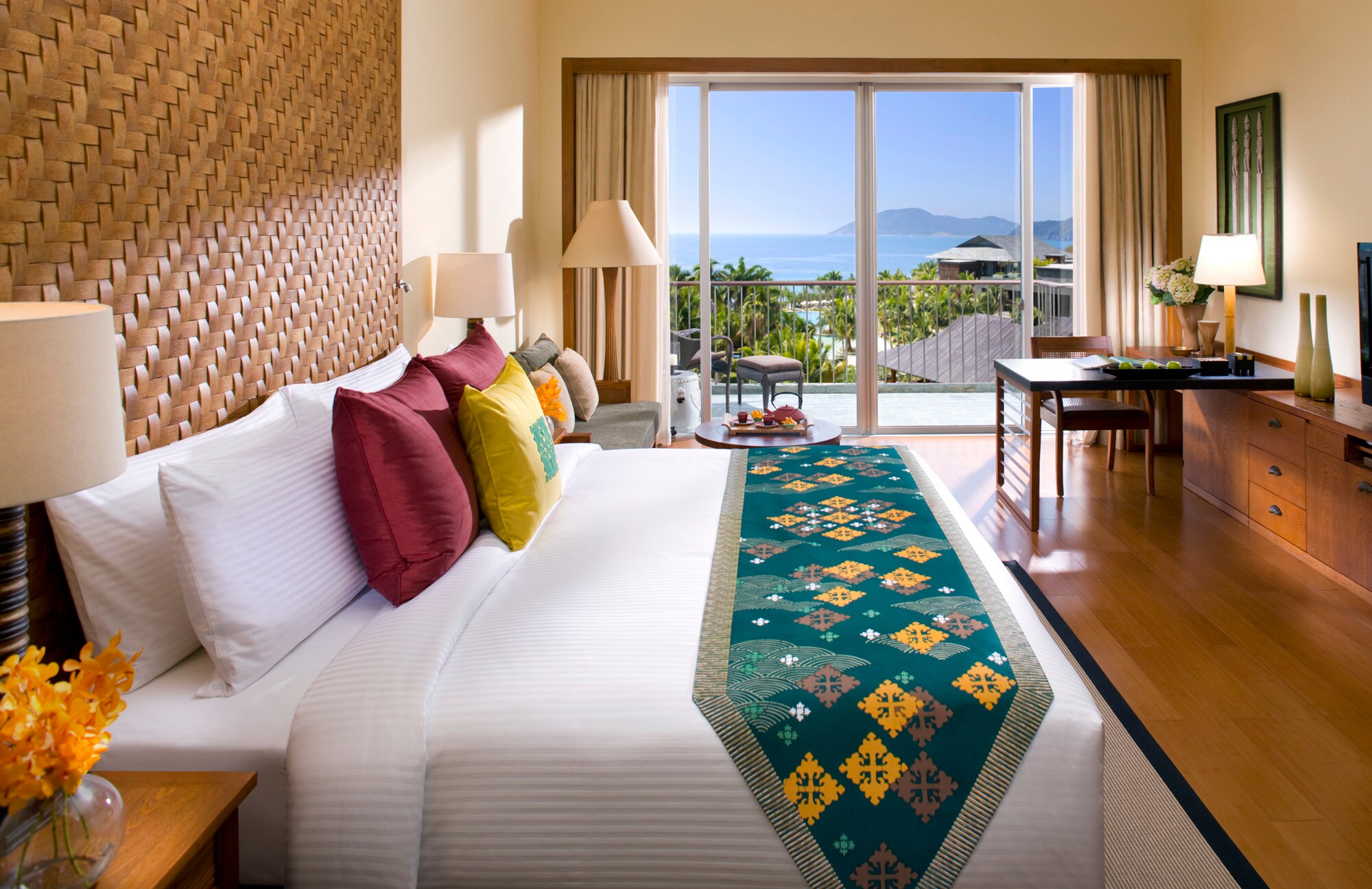 An Ocean View Guest bed is decorated with a wicker headboard and brightly embroidered cushions. Light streams in from glass balcony doors.