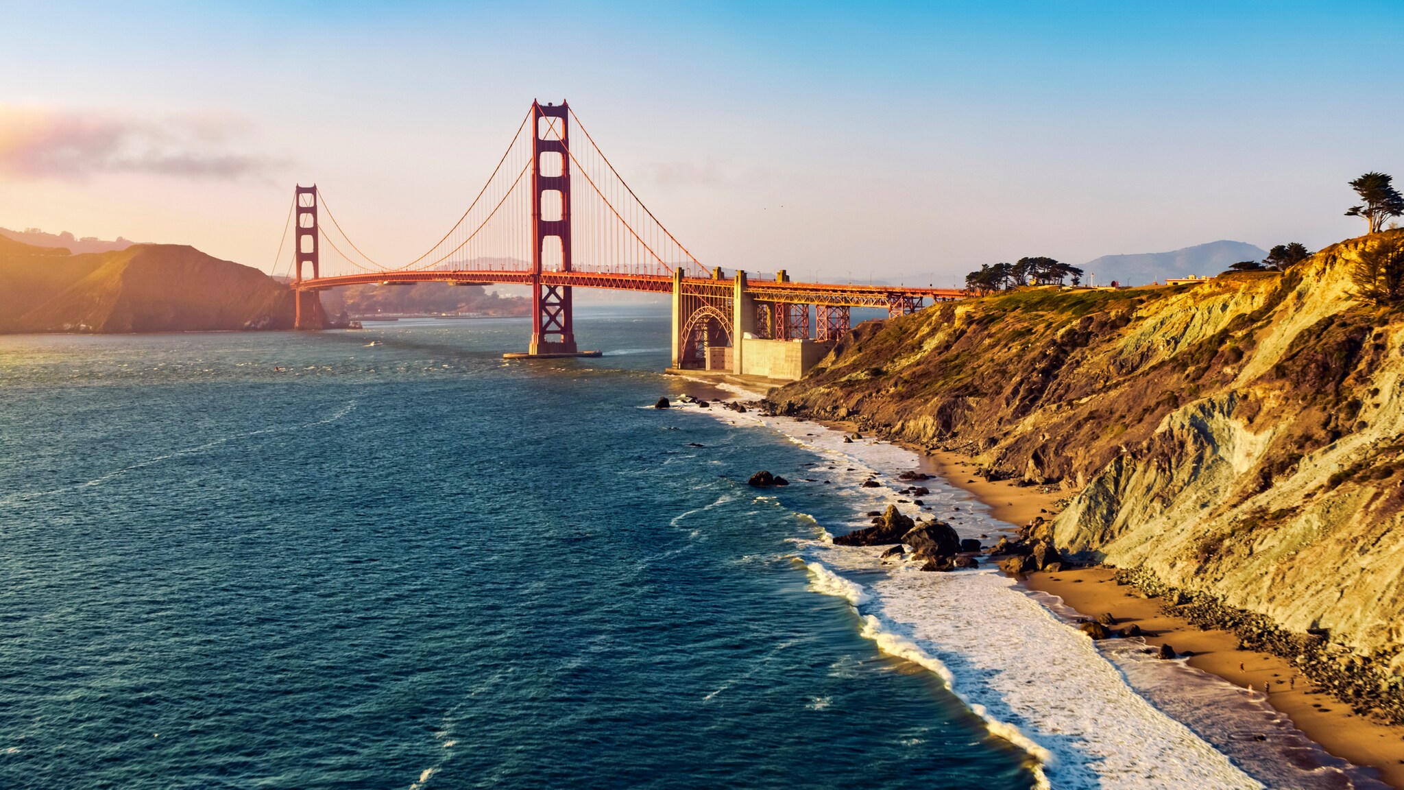 Aerial view of steep hills curving into the Golden Gate Bridge at sunset. The hills slope down to a beach dotted with people.