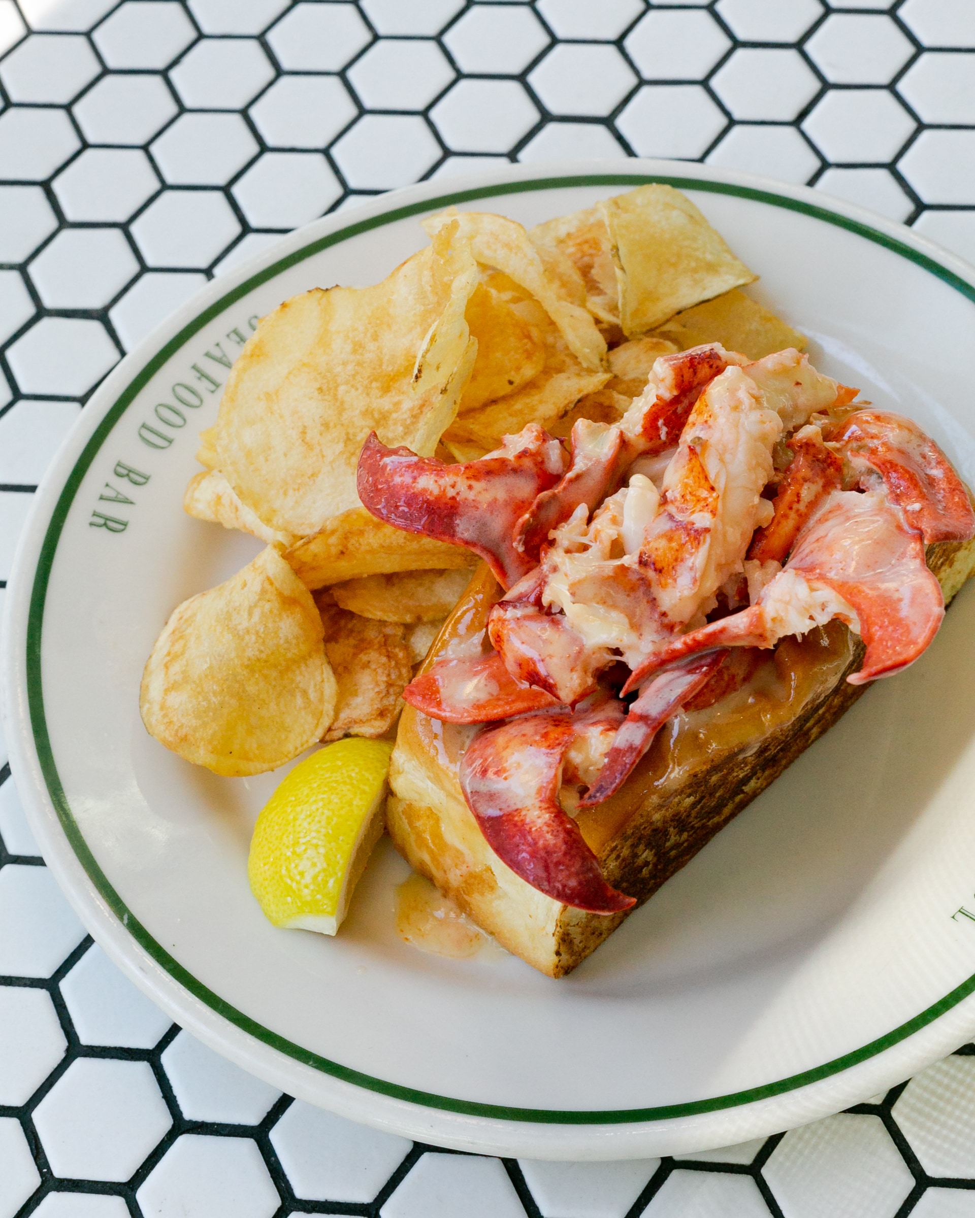 Close-up view of a toasted lobster roll glistening with sauce on a white plate, accompanied by a wedge of lemon and a pile of potato chips.