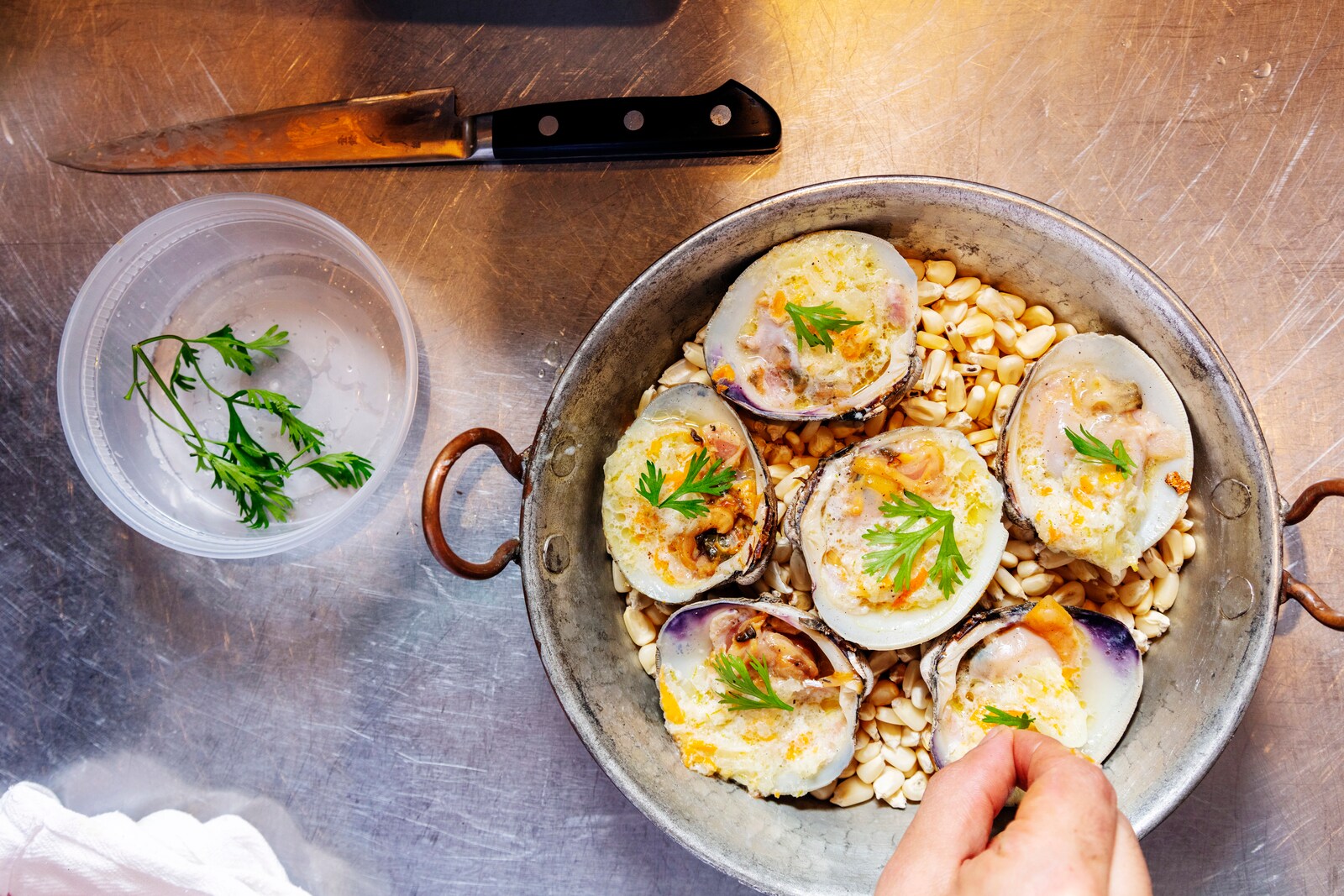 Overhead view of a pot with shellfish nestled on top of corn as a chef garnishes each item with parsley.