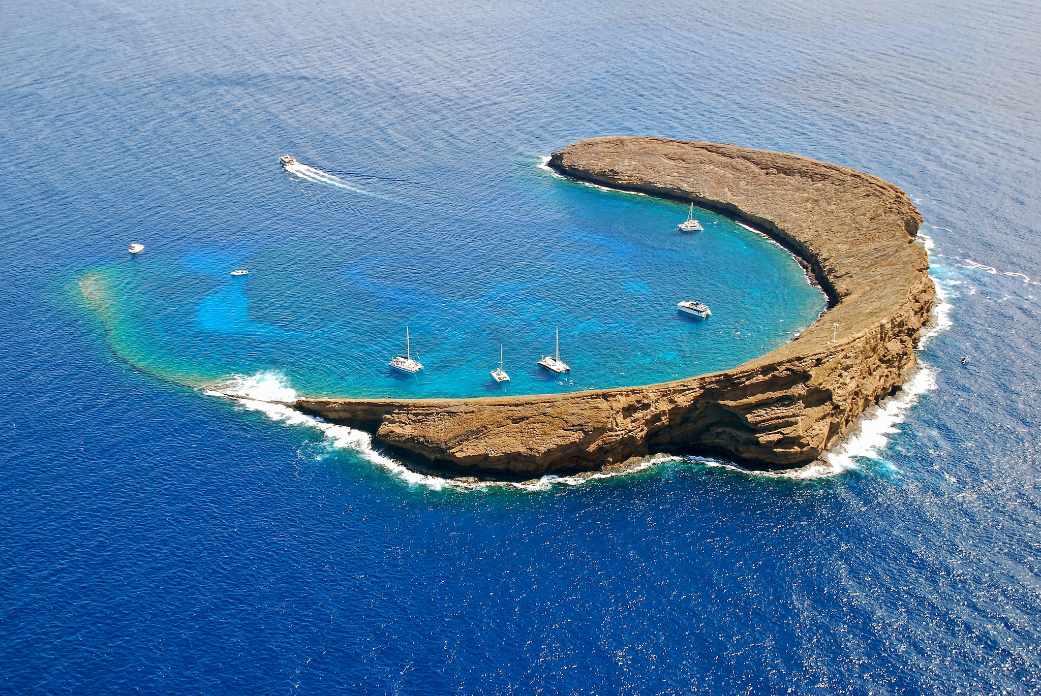 Aerial view of Molokini, a rocky, semicircular islet sheltering several boats in the middle of deep blue water.