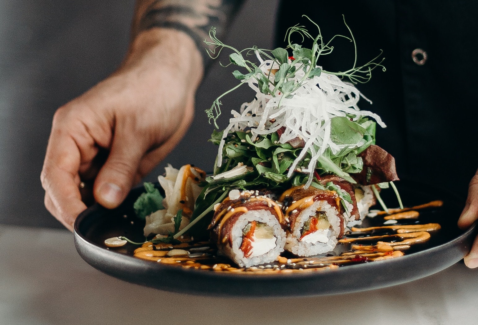 Chef’s hands hold a black dish with sushi rolls covered in lines of sauce, with a pile of greens and sprouts balanced on top of the sushi.
