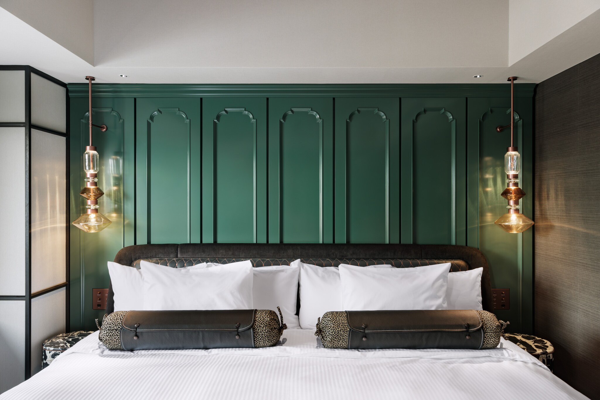 A wide bed with a plush black headboard rests against a green paneled wall, with two industrial art deco lights hanging on either side.