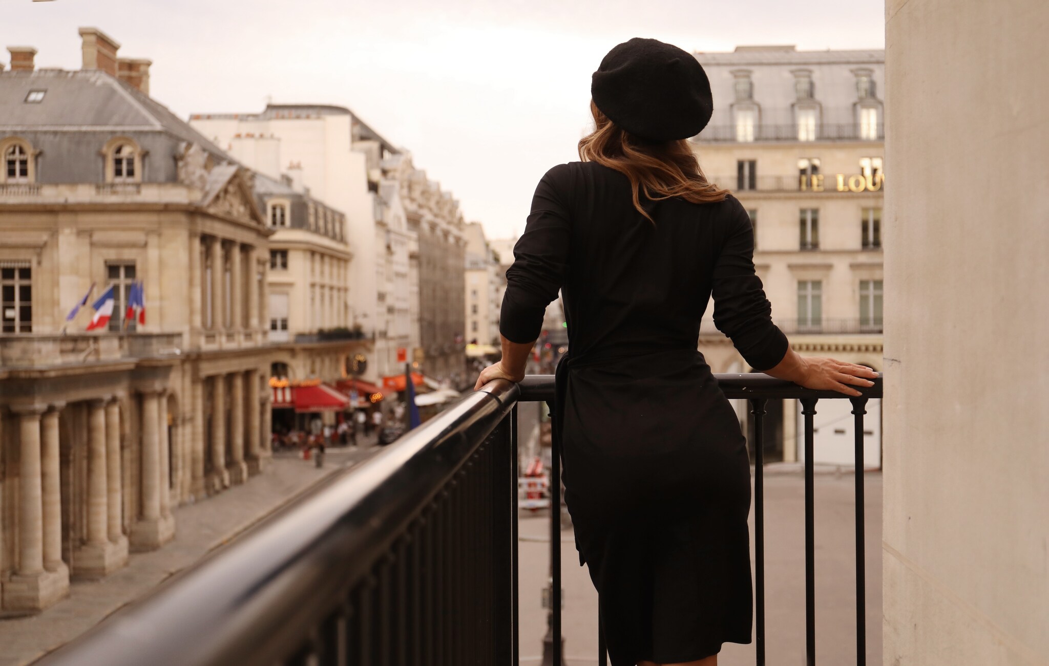 View from behind of a white woman with blonde hair in a black dress wearing a black beret standing on a balcony looking out at the street.