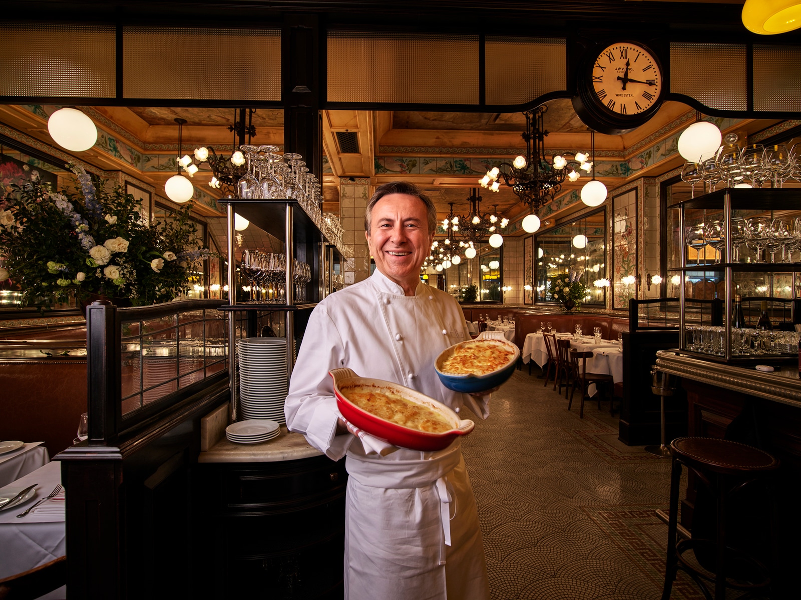 Chef Daniel Boulud holds out two casserole dishes as the dining room of Le Gratin stretches out behind him.