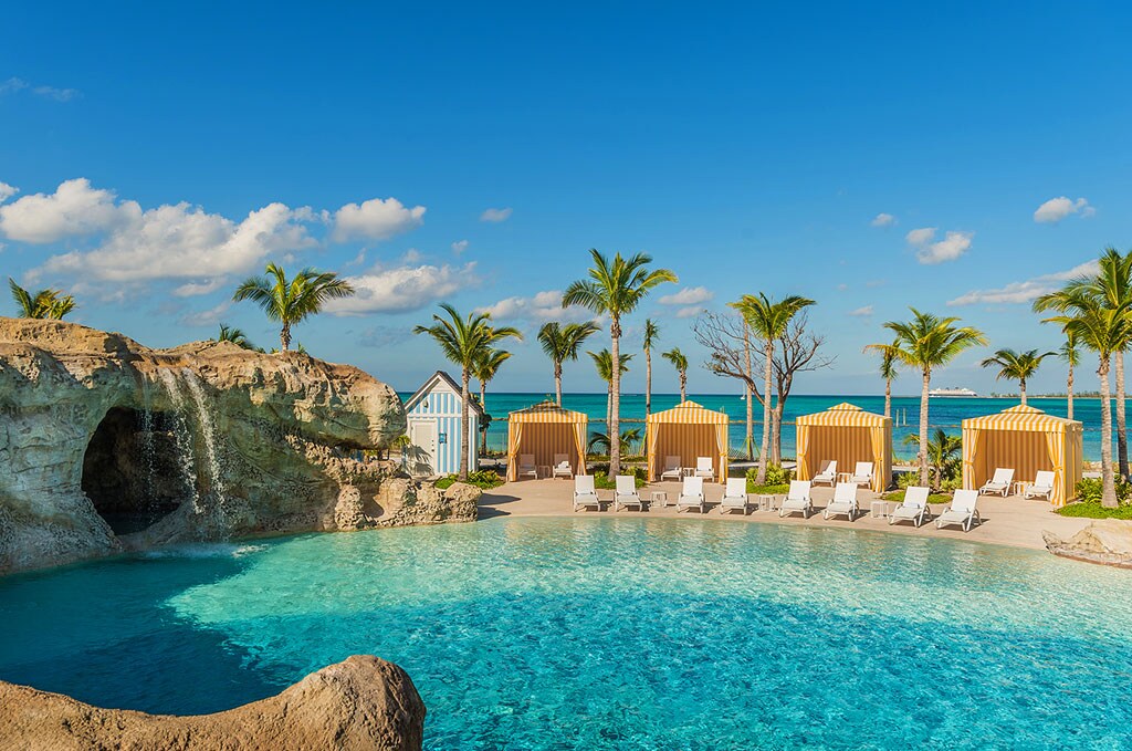 Water arcs off an ochre cliff into a large blue pool. Sun loungers and cabanas line the deck, with palm trees and the beach visible beyond.