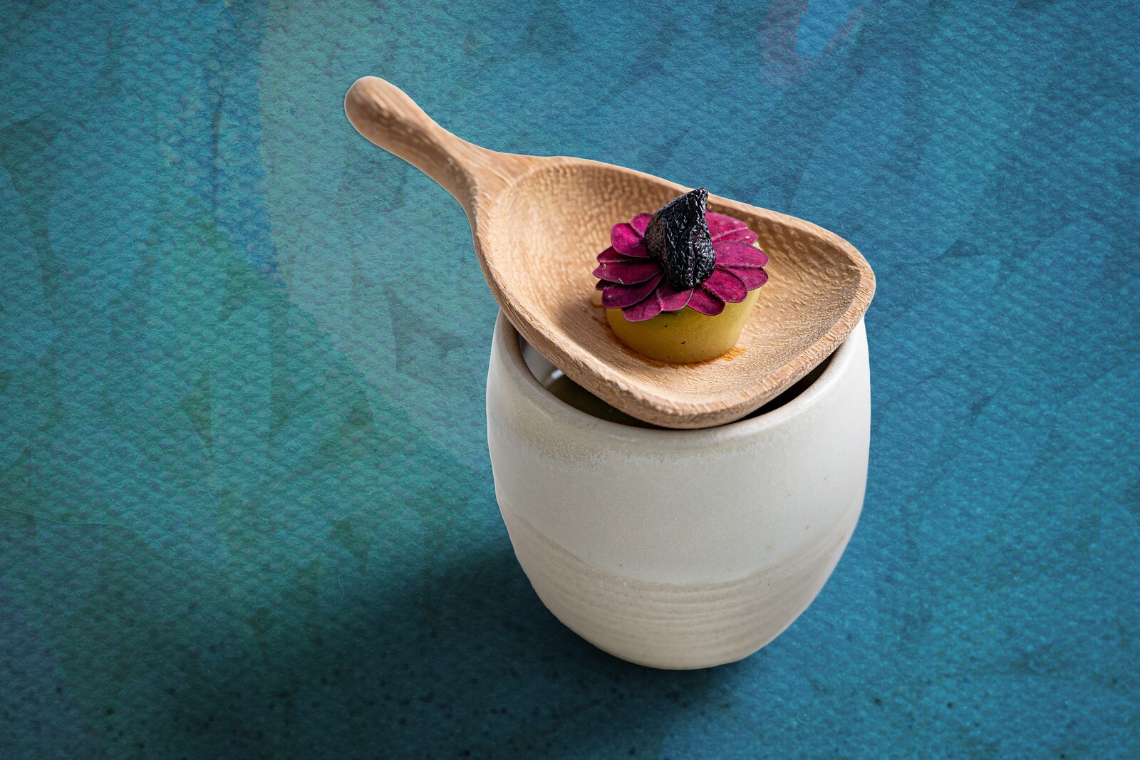 A wide wooden spoon is balanced on a ceramic cup of liquid, holding a multicolored flower of food.