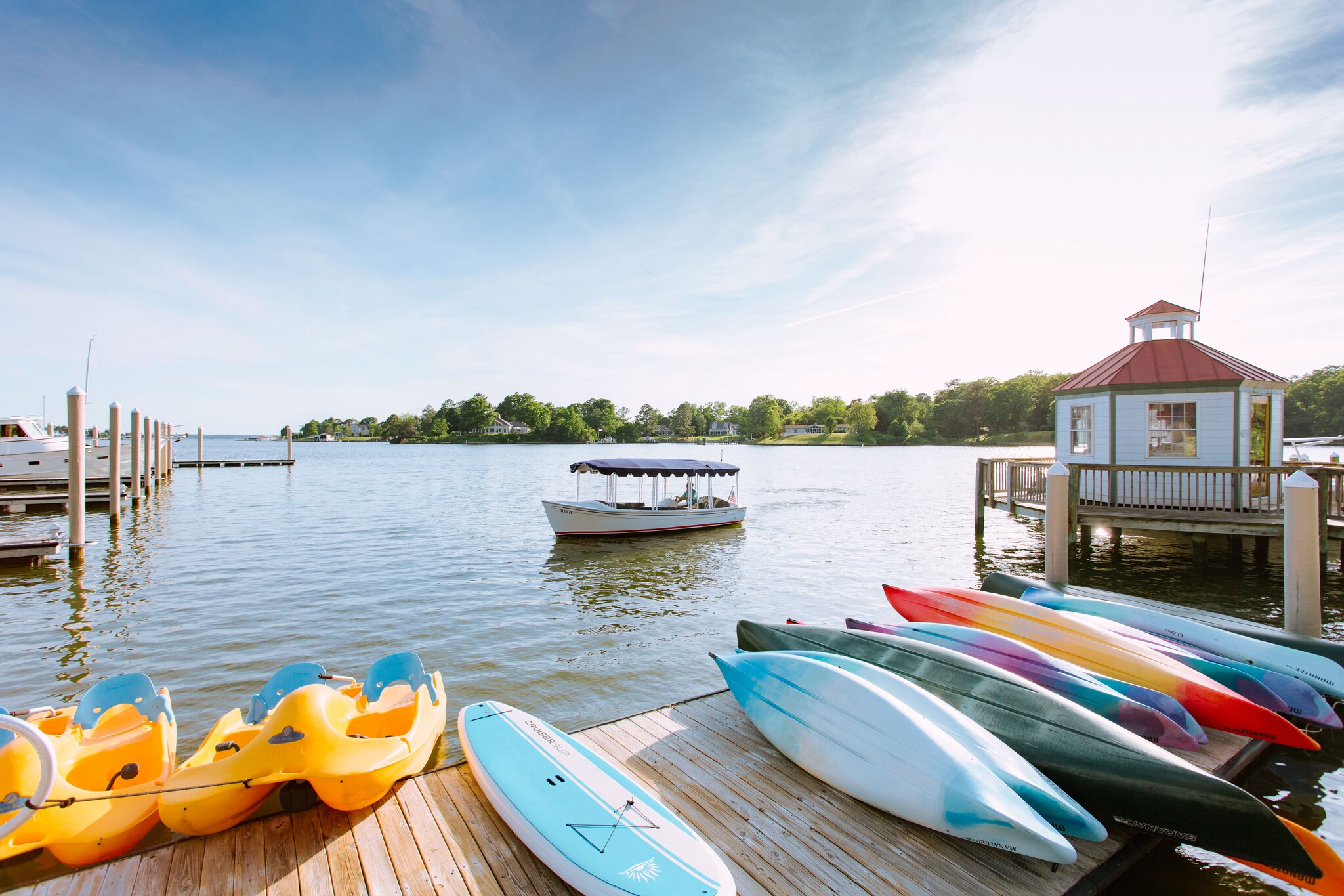 On a dock on a sunny day, kayaks and paddleboards are stacked next to each other, with more docks and boats visible in the background.