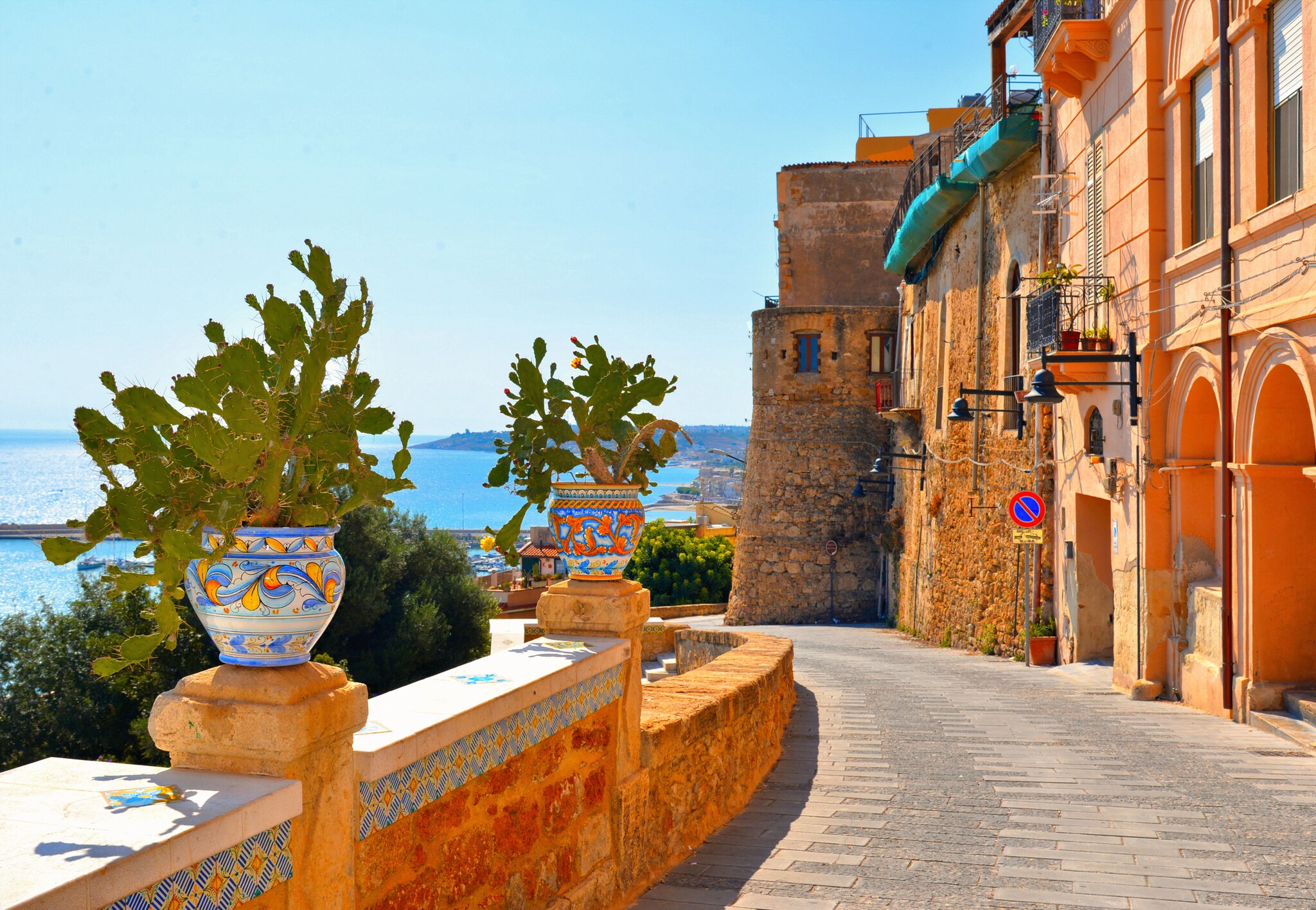 A sunny street near the waterfront. The buildings are built of pastel orange stone, and richly painted vases punctuate the roadside wall.