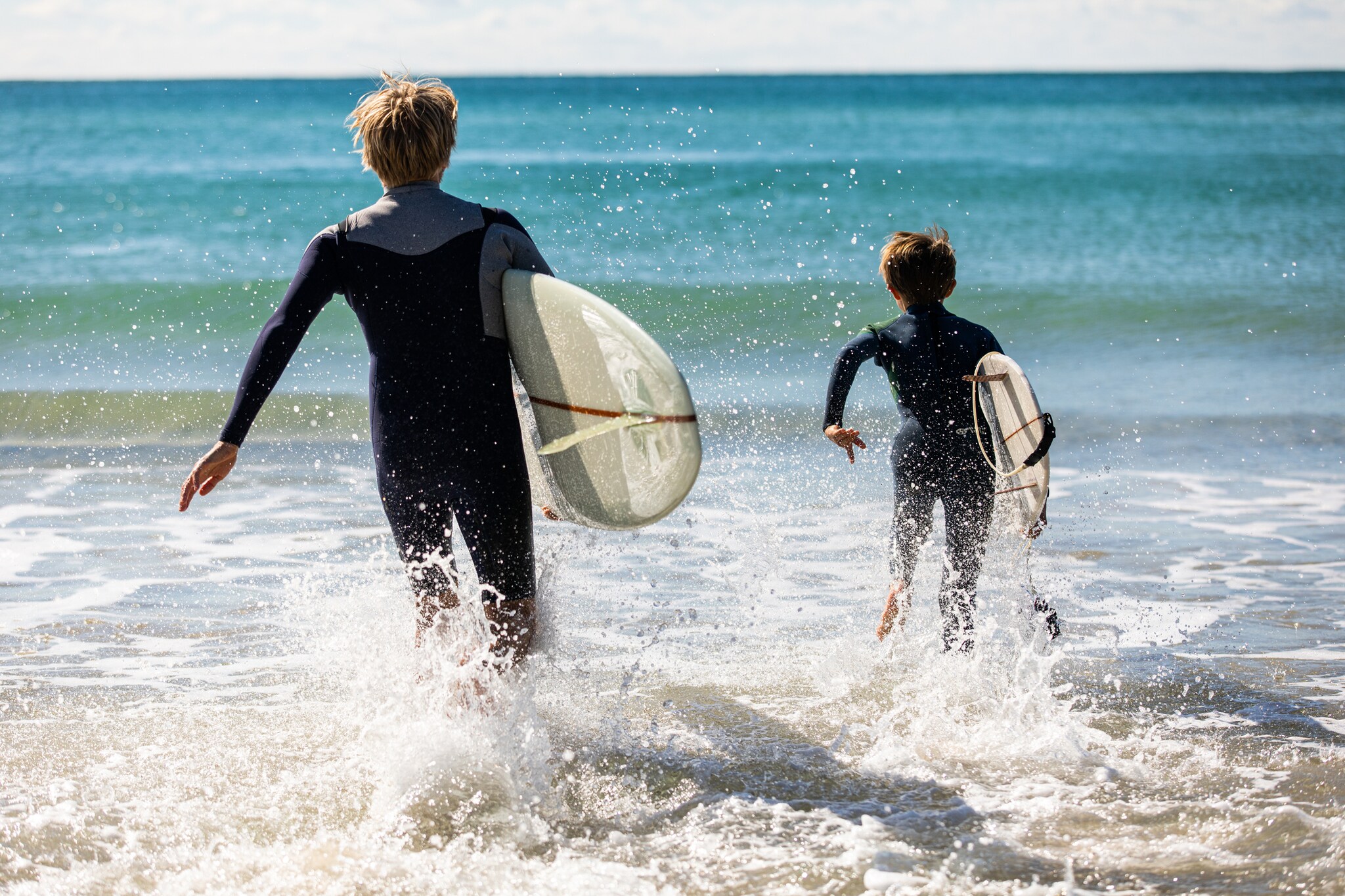 A white man with short blond hair and a white boy with brown hair run in wetsuits, with surfboards under their arms, into the ocean.