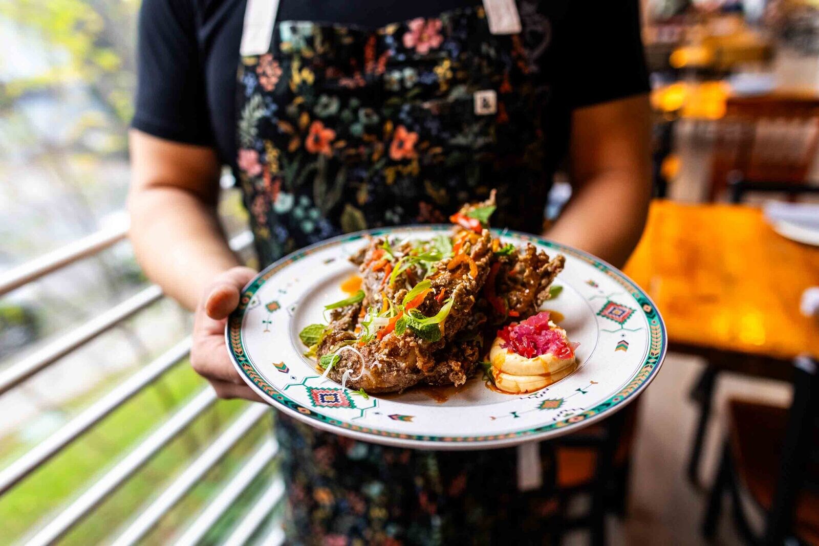 A chef in a floral apron holds out a plate of food. The dish is a pile of crispy meat dotted with sprigs of herbs and vegetables.