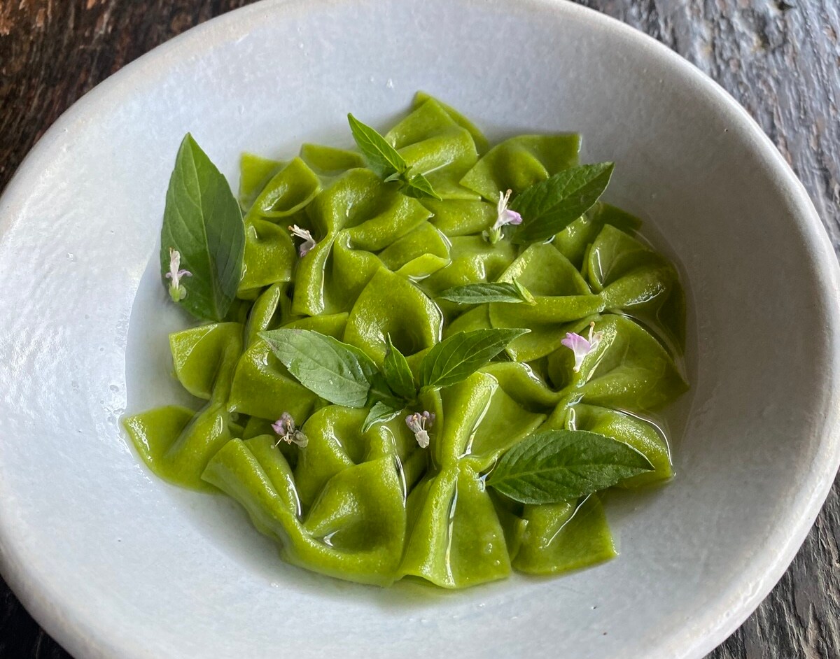 A bowl of green bowtie pasta rests in a clear broth with garnishes of basil leaves and small pink flowers.