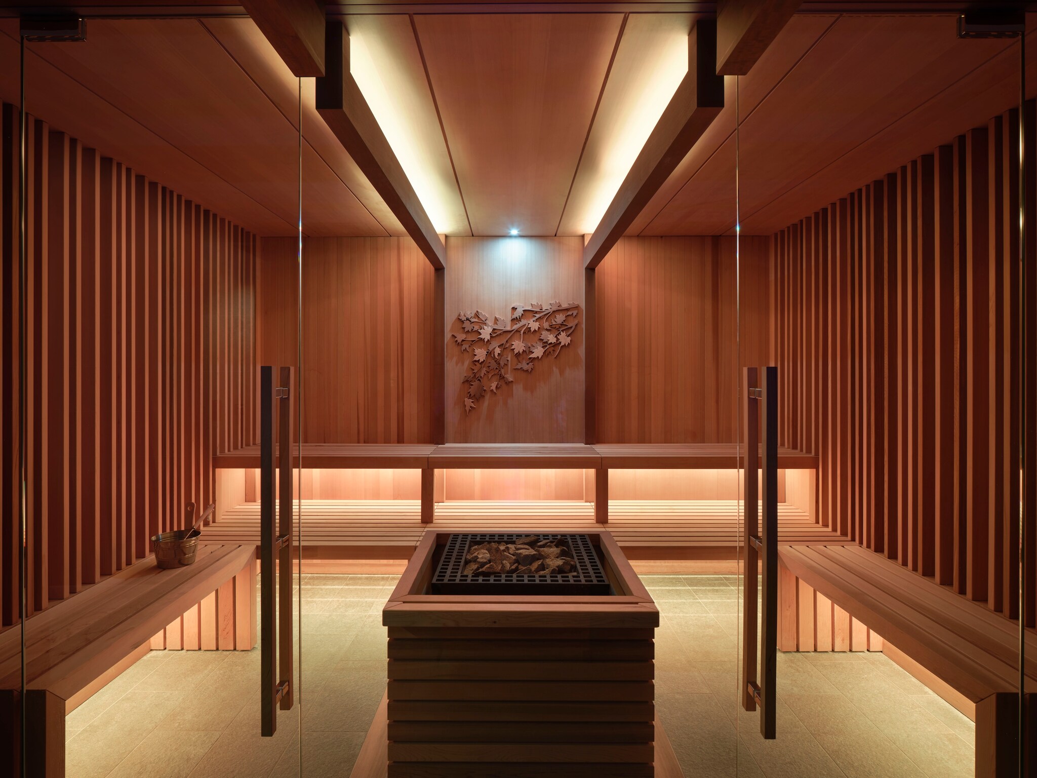 Interior of a sauna. Vertical wood panels line the walls and wooden benches offer seating around a square heater stacked with wood chips.