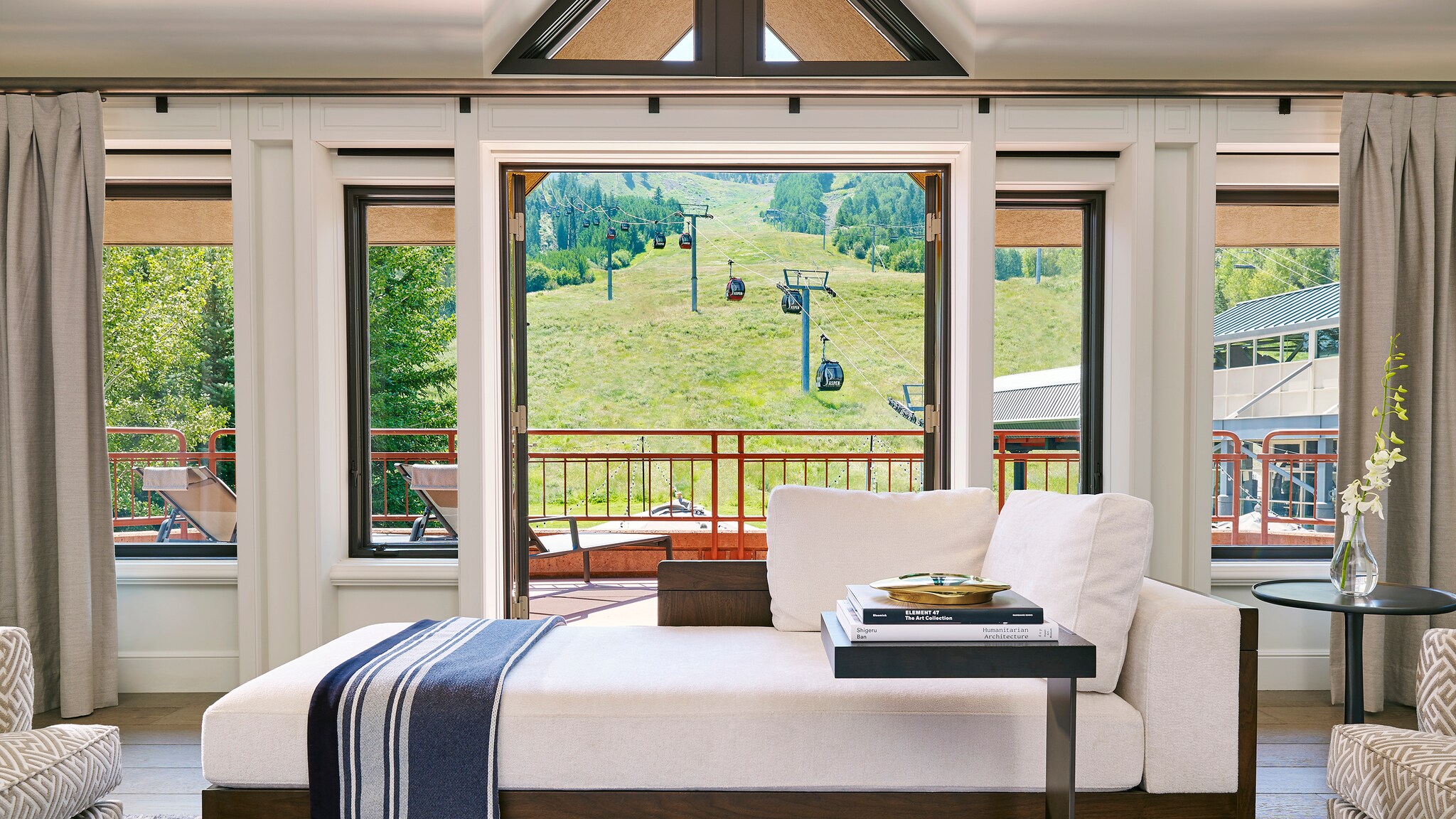 In the Paepcke Suite, a white day bed looks out glass doors to a patio with lounge chairs. Beyond, a green slope is traversed by a funicular.