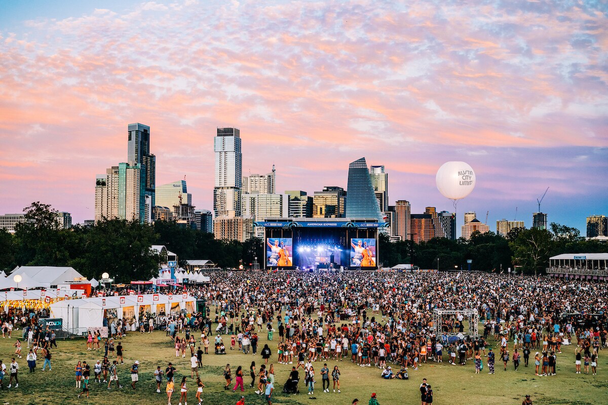 An aerial view of Austin City limits captures the crowds filling a wide field in front of a stage, which is backed by the city skyline.
