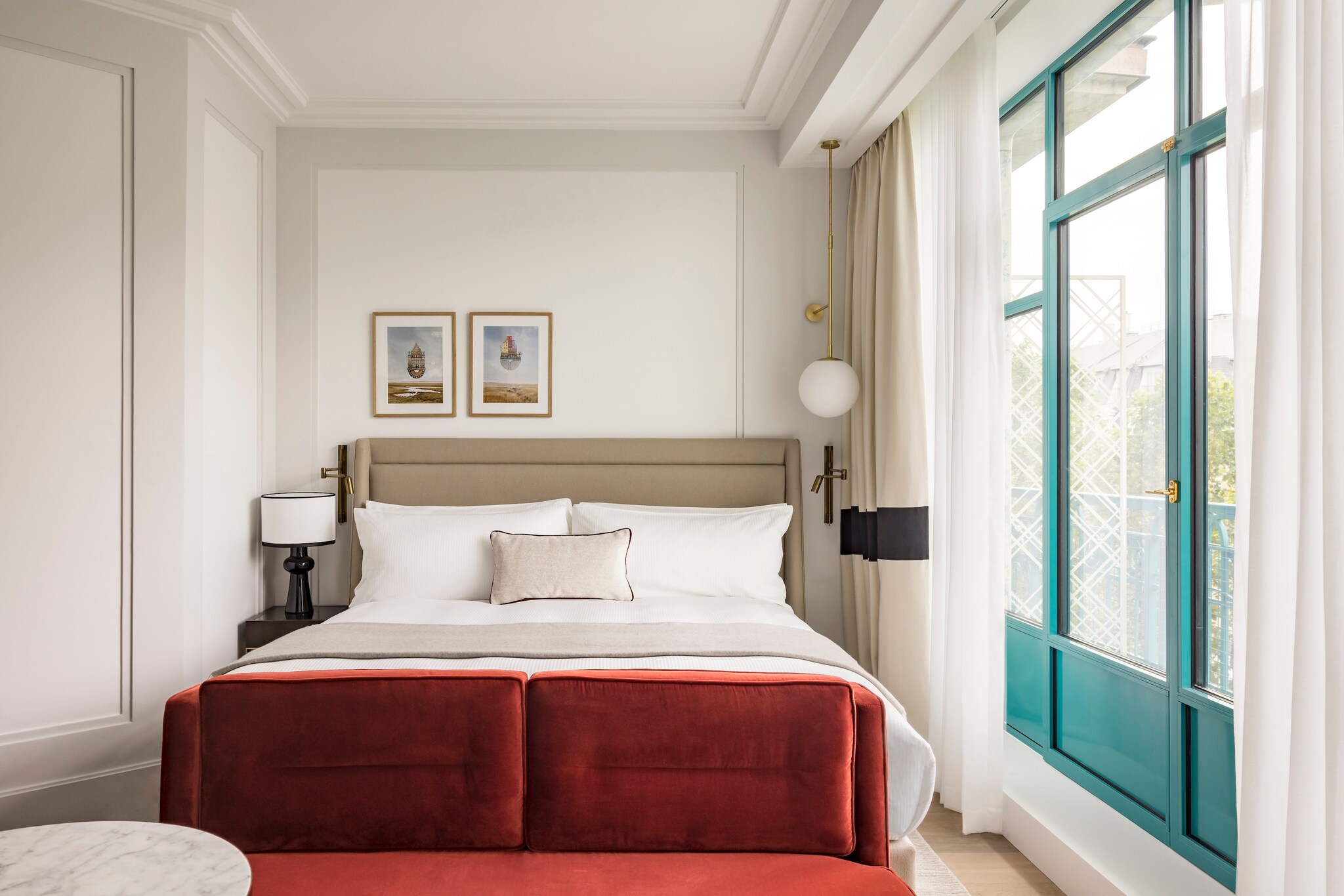 In a hotel room, a red loveseat is situated at the bottom of the bed, and a glass door with a bright turquoise frame opens into a balcony.