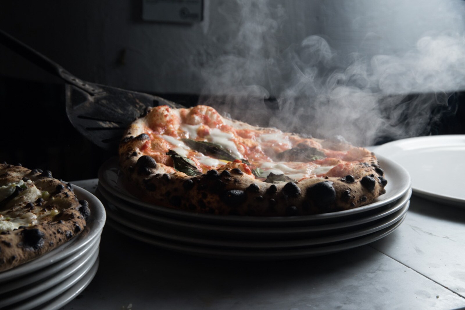In a dark room, a steaming Neopolitan-inspired margherita pizza slides onto the top of a stack of white plates.