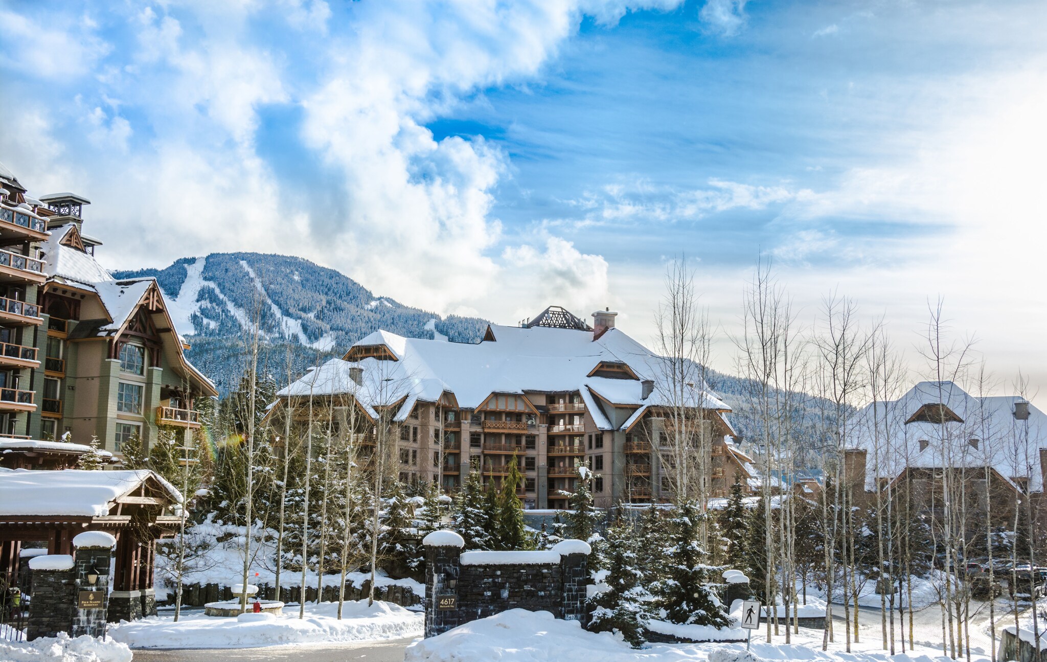 Exterior view of the property, with several multi-story buildings arrayed around birch and pine trees, and snowy mountains beyond.