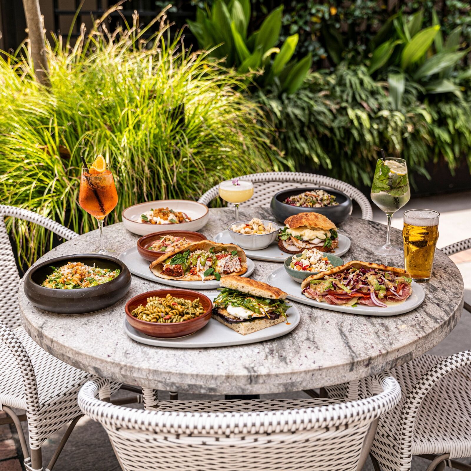 A round marble table is laden with dishes filled with pasta, salad, paninis, and pita. Beer and cocktails are scattered throughout.