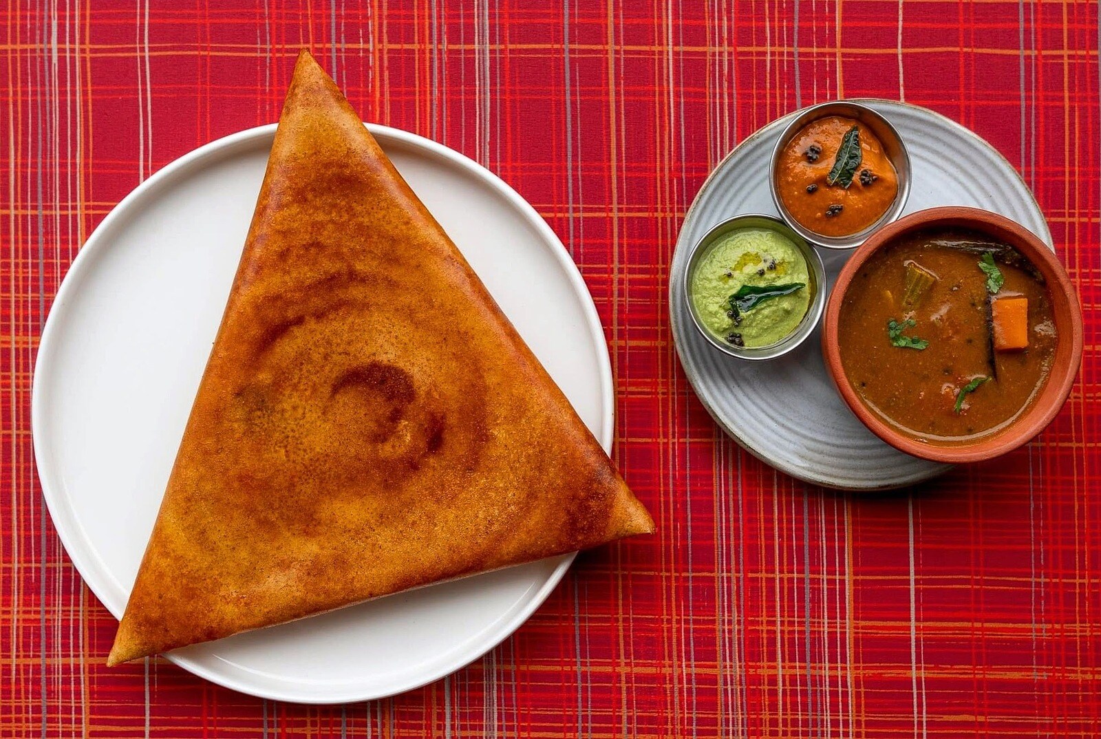 A massive dosa folded into a huge triangle pokes over the side of a circular white dish, with a plate of chutneys nearby.