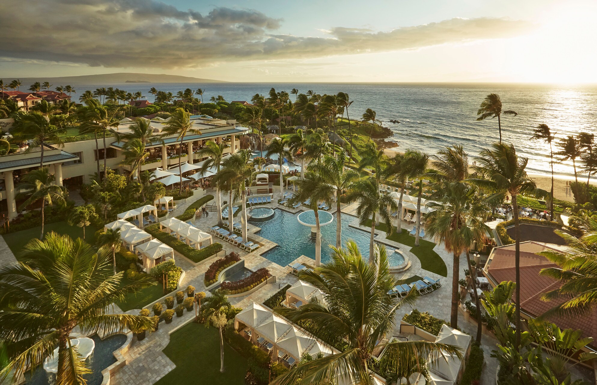 Aerial view of Four Seasons Resort Maui at Wailea. Palm trees bend slightly in the wind around a cross-shaped pool and lines of cabanas.