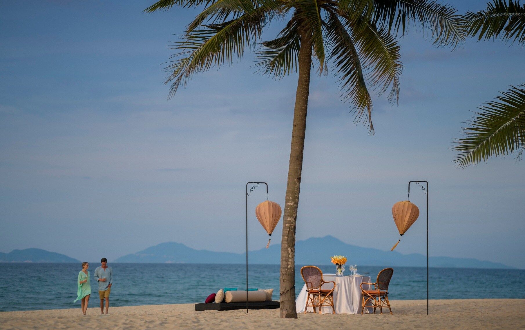A couple walks on the beach towards a table set for two under a palm tree and two hanging lantern stands.