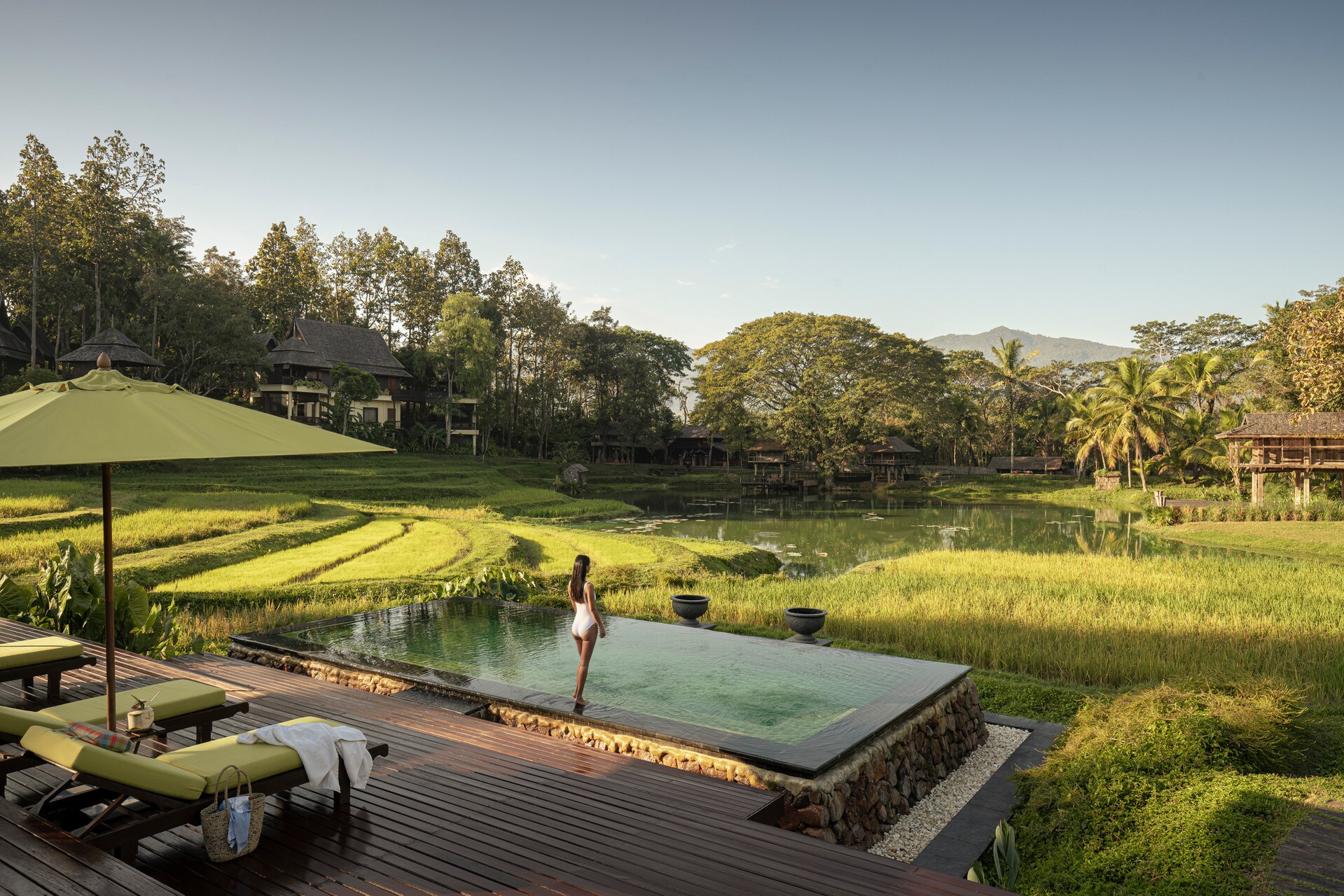 On a terraced hillside, a woman with long, dark hair in a white swimsuit steps into an infinity pool next to a deck set with green sun beds.