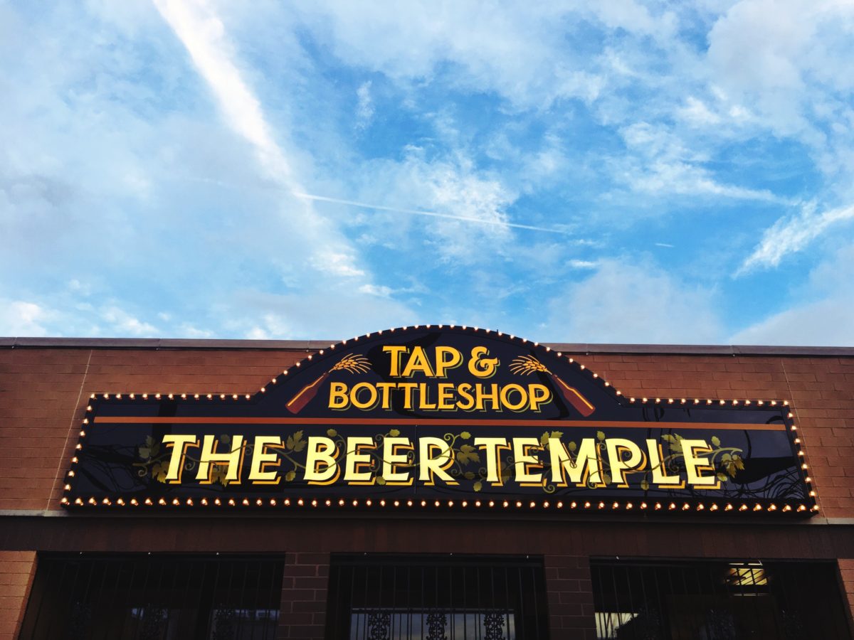 A sign above the store reads “Tap & Bottleshop – The Beer Temple” with hop vines growing through the letters and wheat stalks framing them.